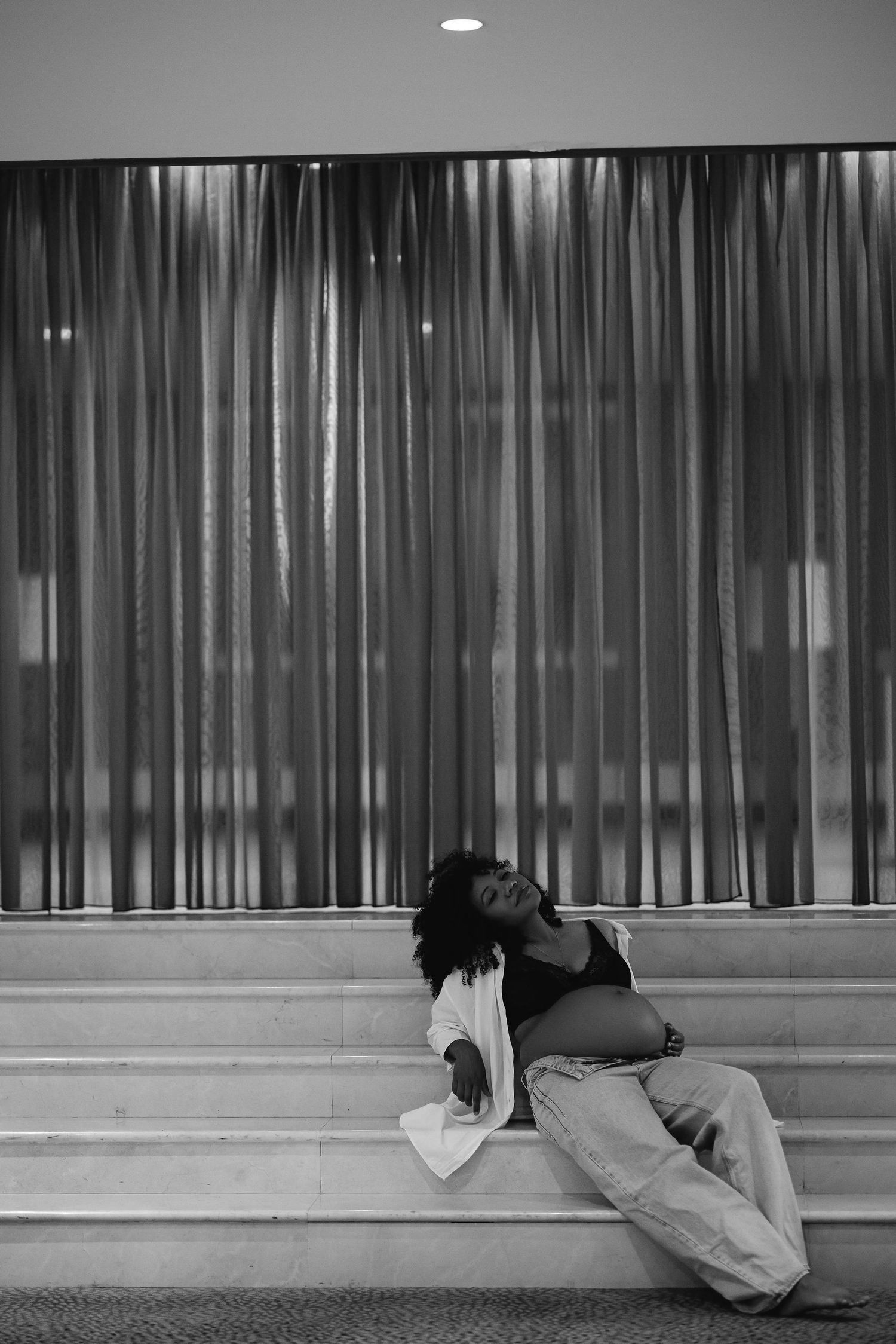 Figure reclining on steps against sheer curtain backdrop in monochrome.