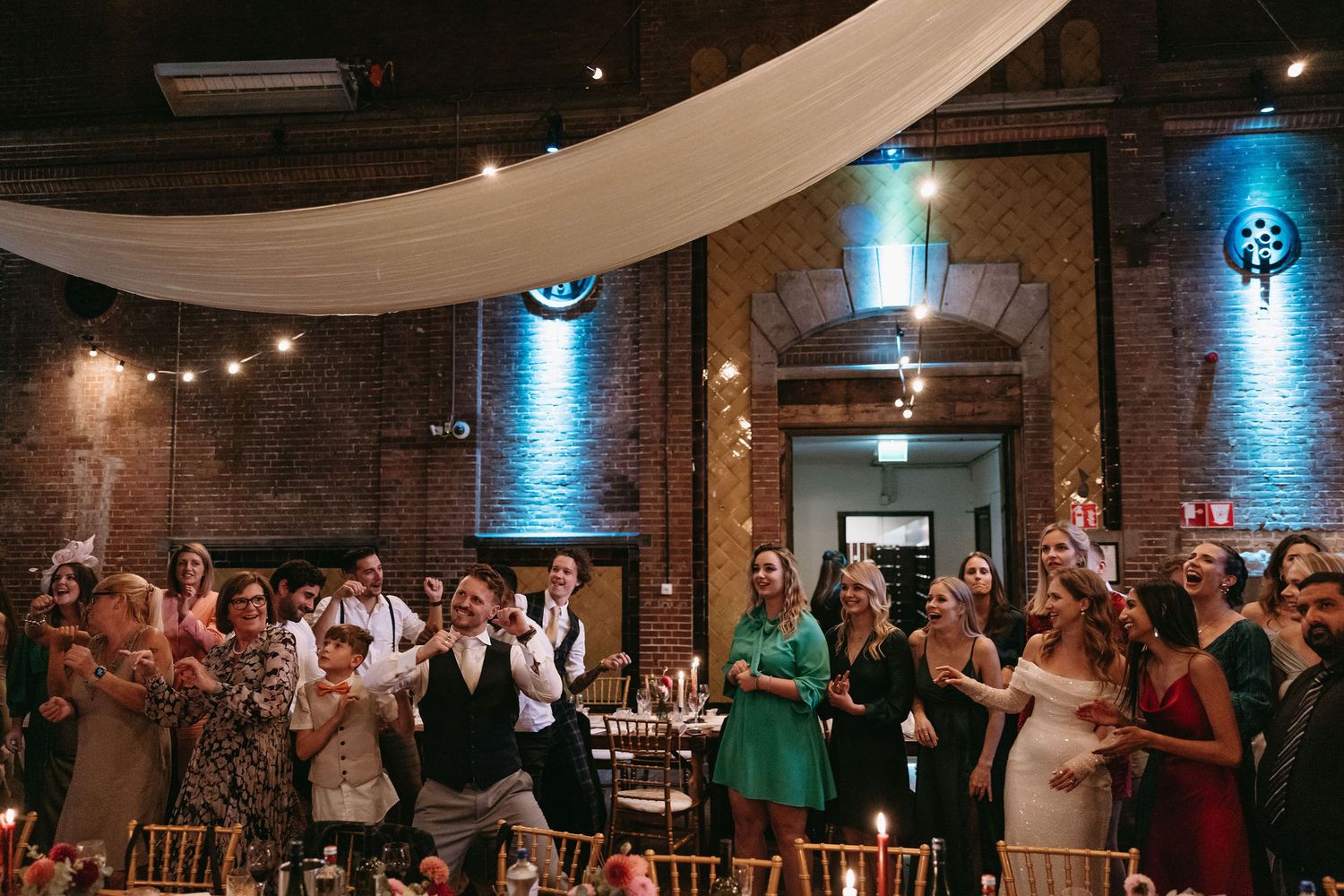Wedding reception venue with exposed brick walls, blue uplighting, and white ceiling draping.