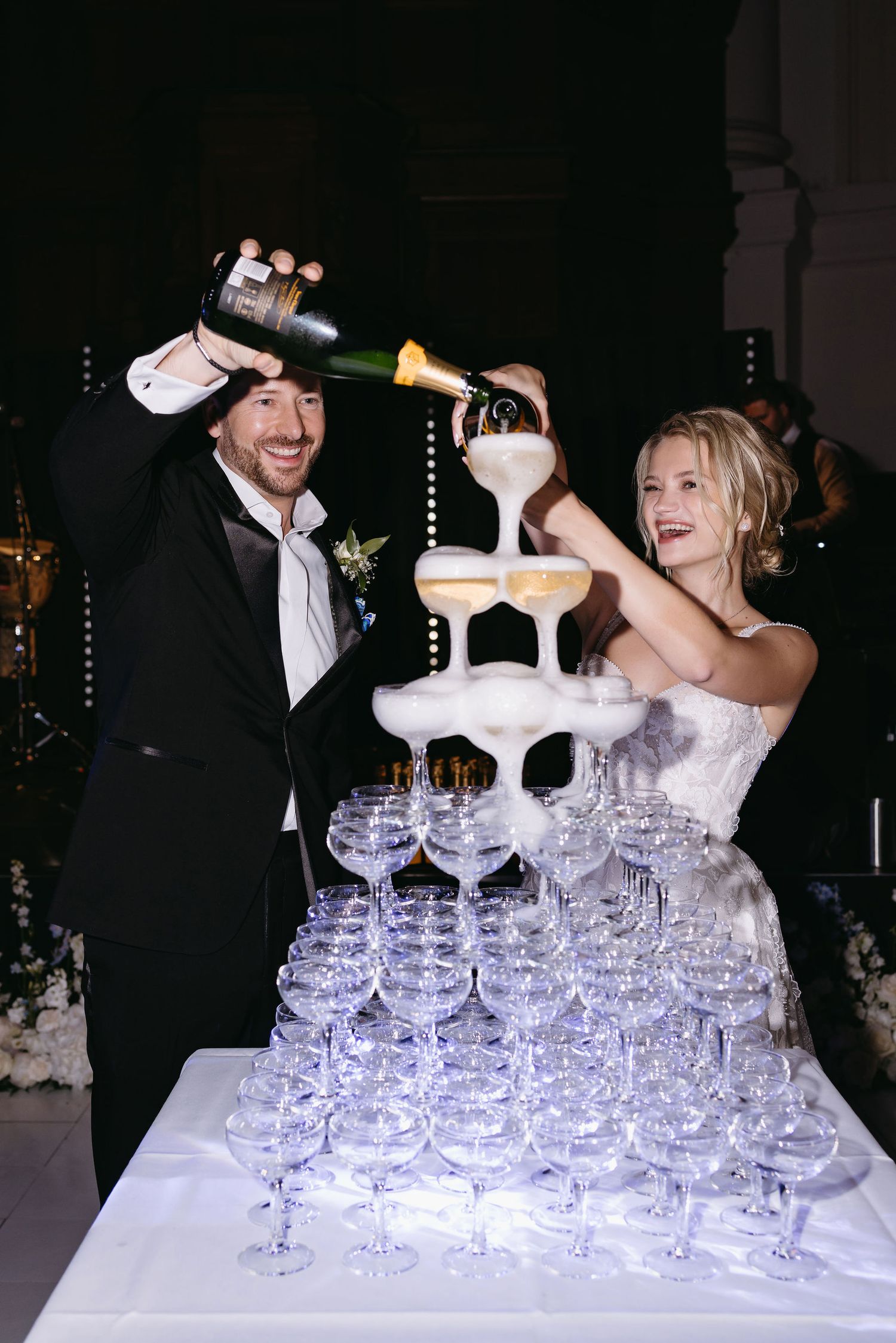 Newlyweds pour champagne into tower of crystal glasses during wedding celebration.