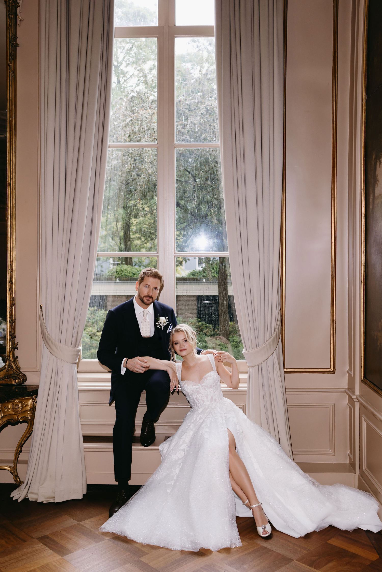 Newlyweds pose dramatically by tall window in elegant ballroom setting.