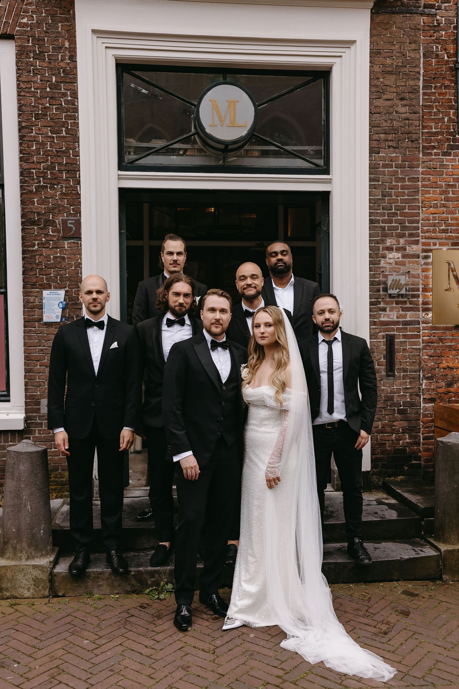 A wedding party poses on brick steps in front of a historic building entrance.