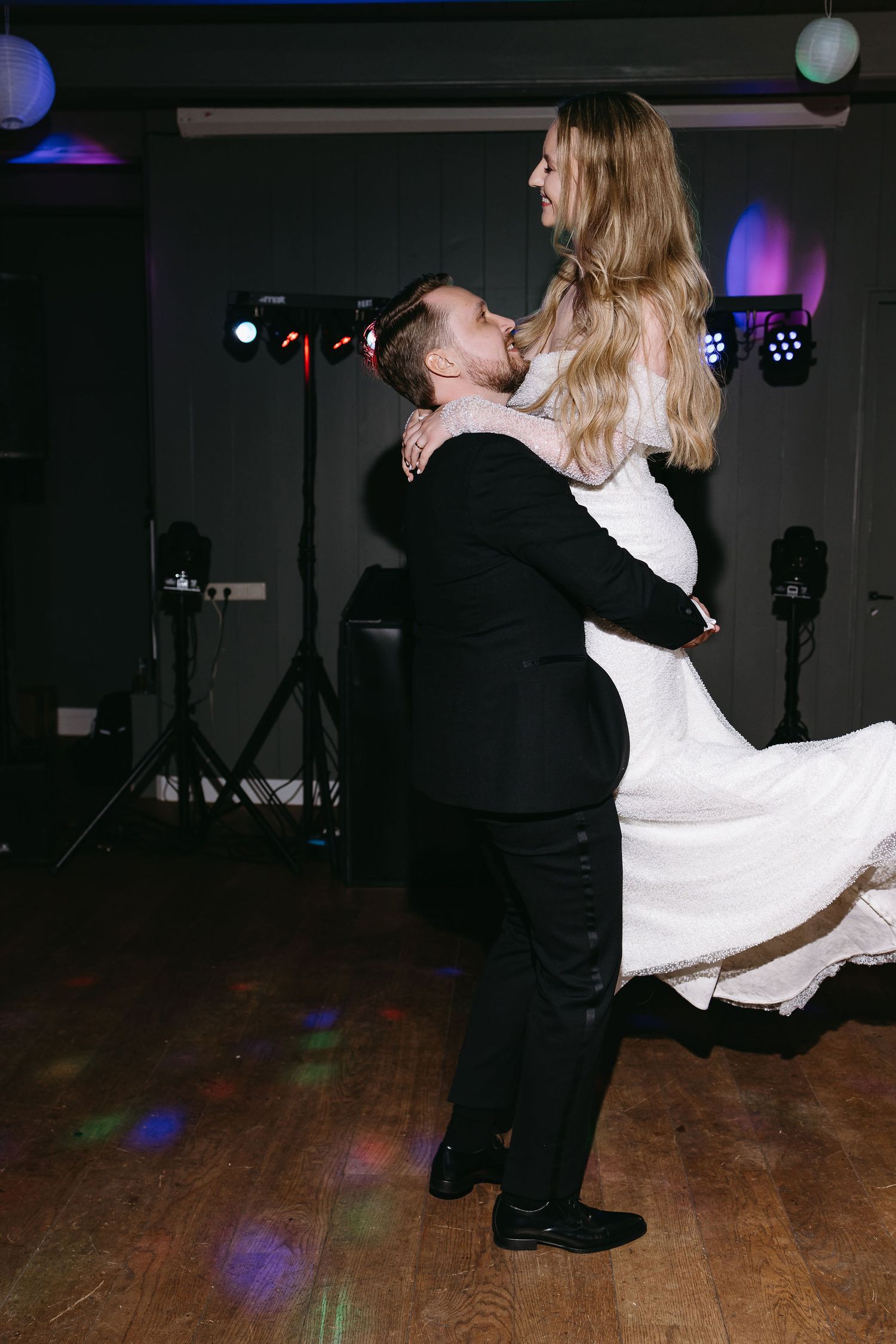 A romantic dance lift moment at a wedding reception with colorful lighting and a wooden dance floor.