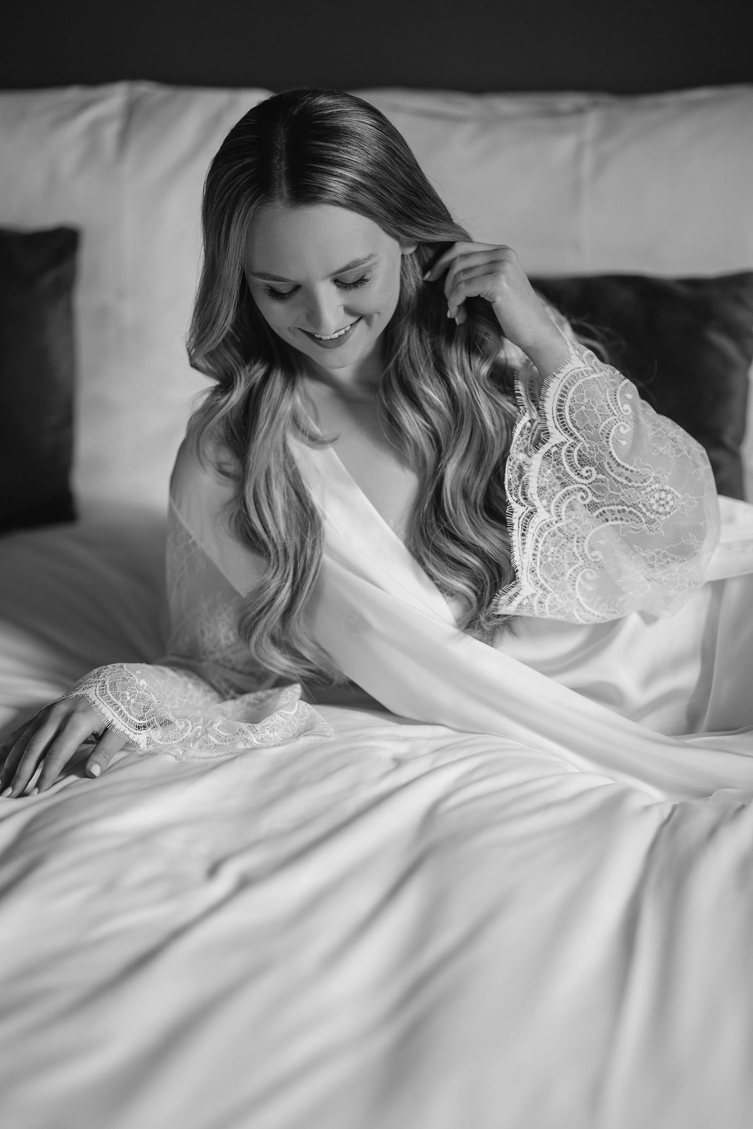 Black and white boudoir photo of woman in silk robe with lace details on white bedding.