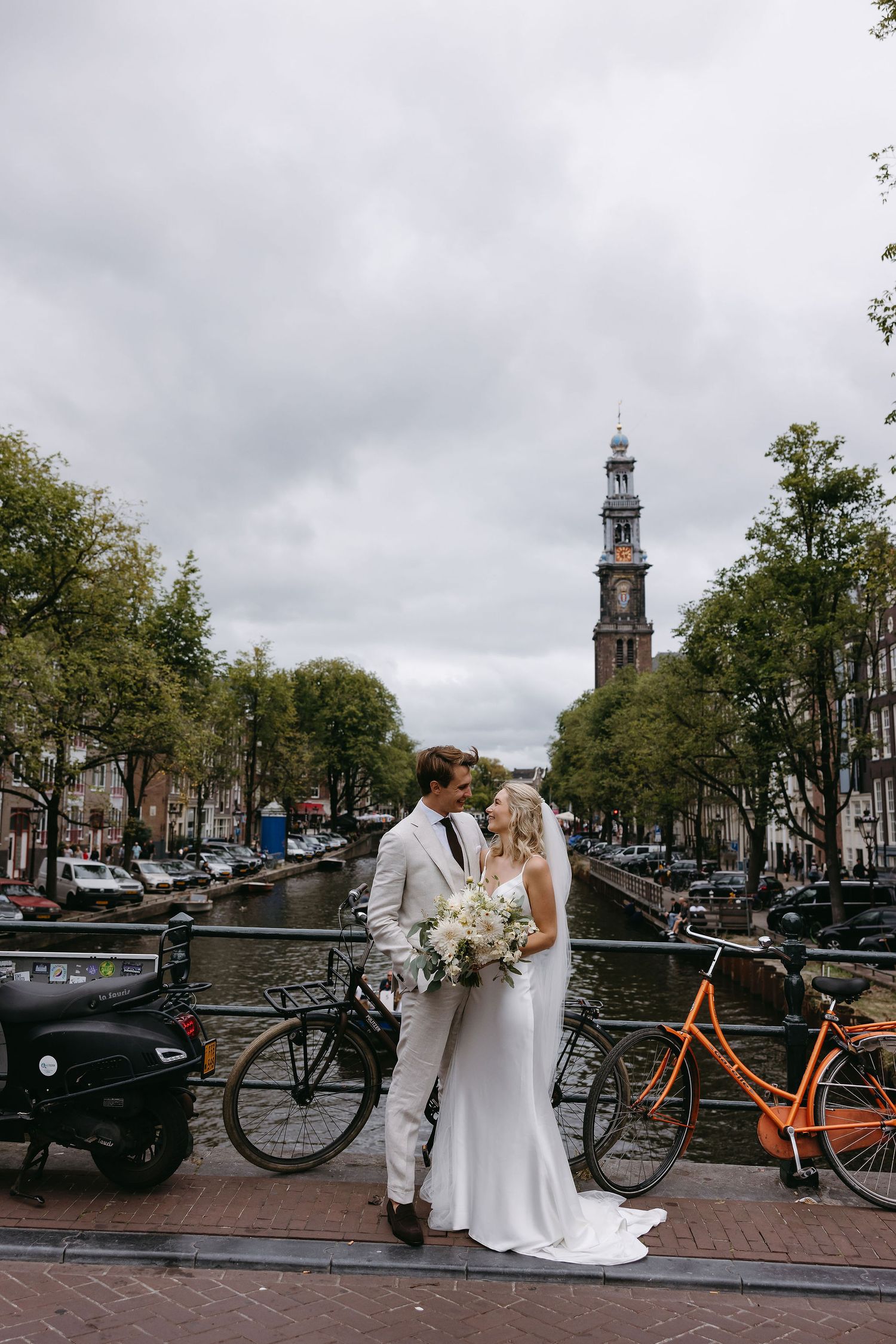 Wedding couple posing by Amsterdam canal with iconic church tower and bicycles in background.
