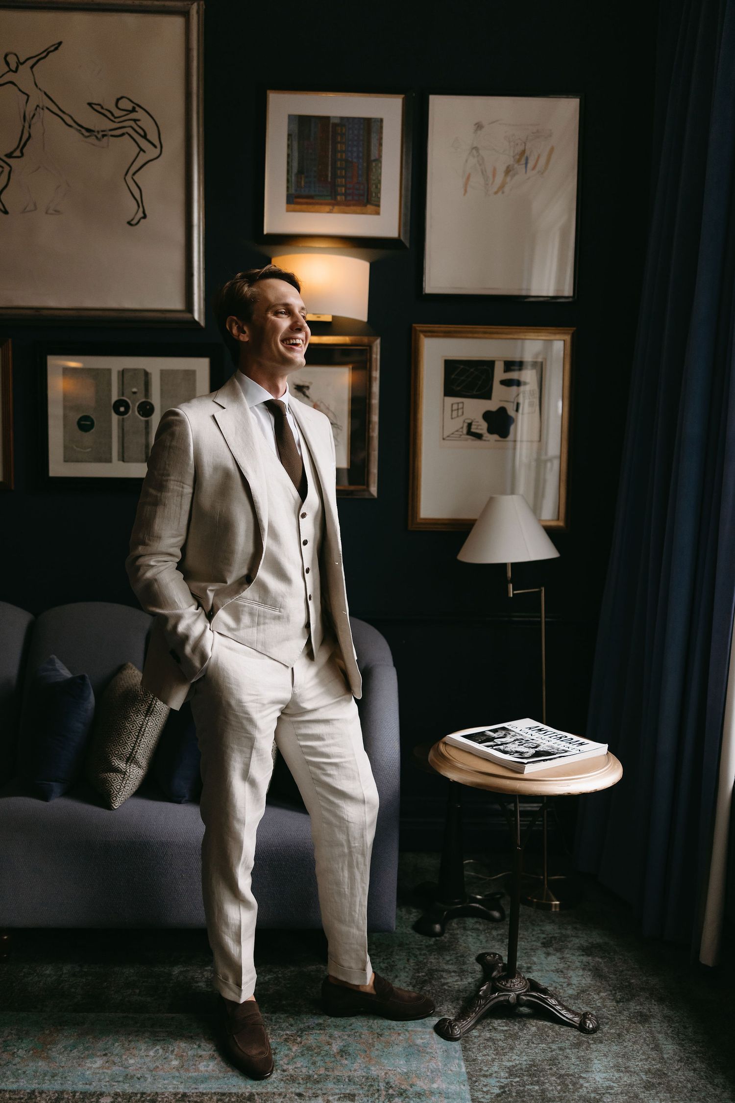 Groom in light beige suit standing in moody room with framed artwork and lamp.