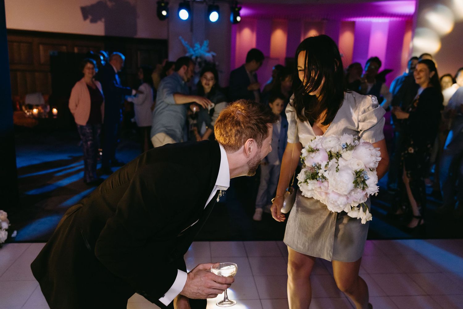 Wedding guests dance and socialize on a dimly lit dance floor with purple lighting effects.