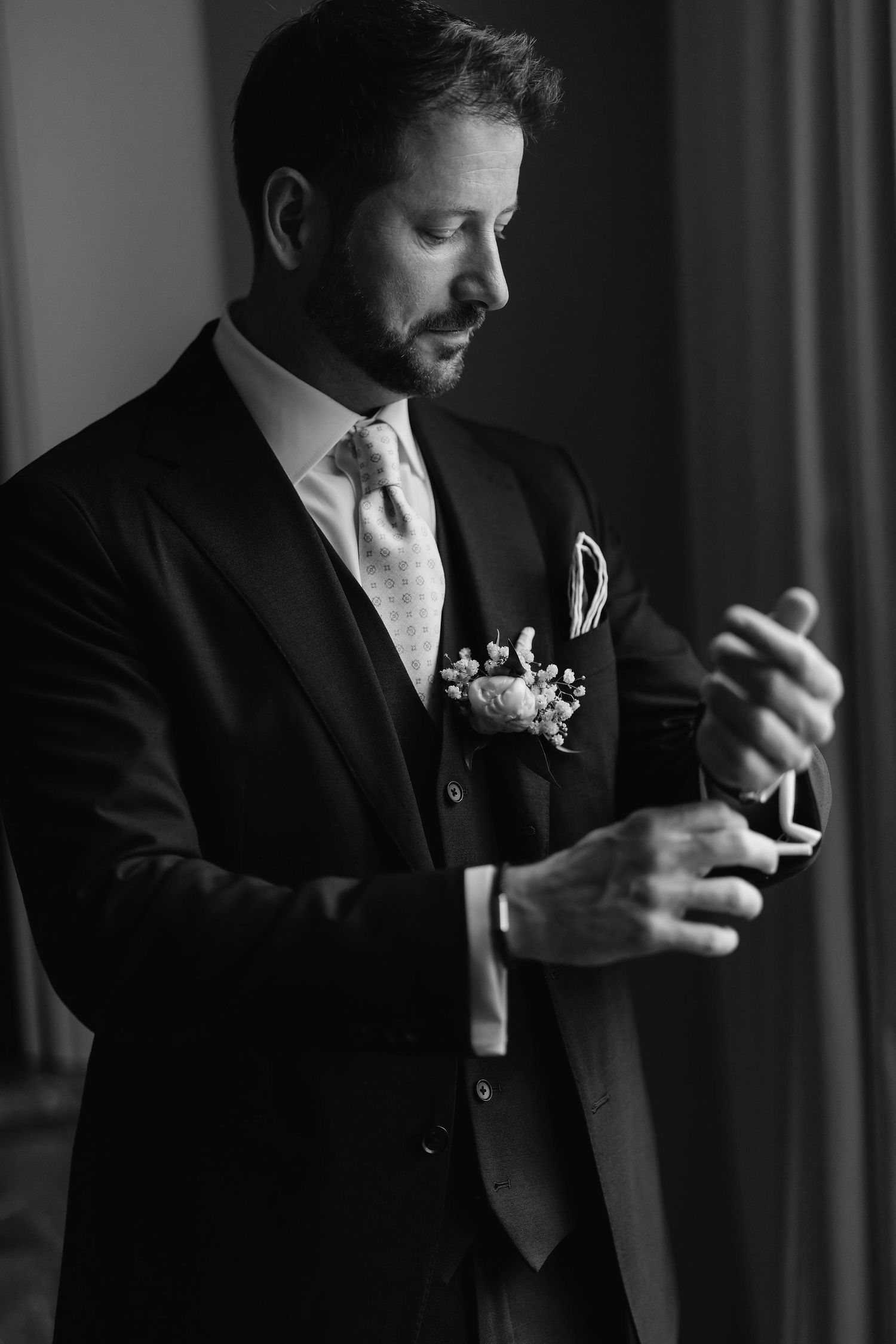 A groom in a classic black suit and tie adjusts his cuff while getting ready on his wedding day.