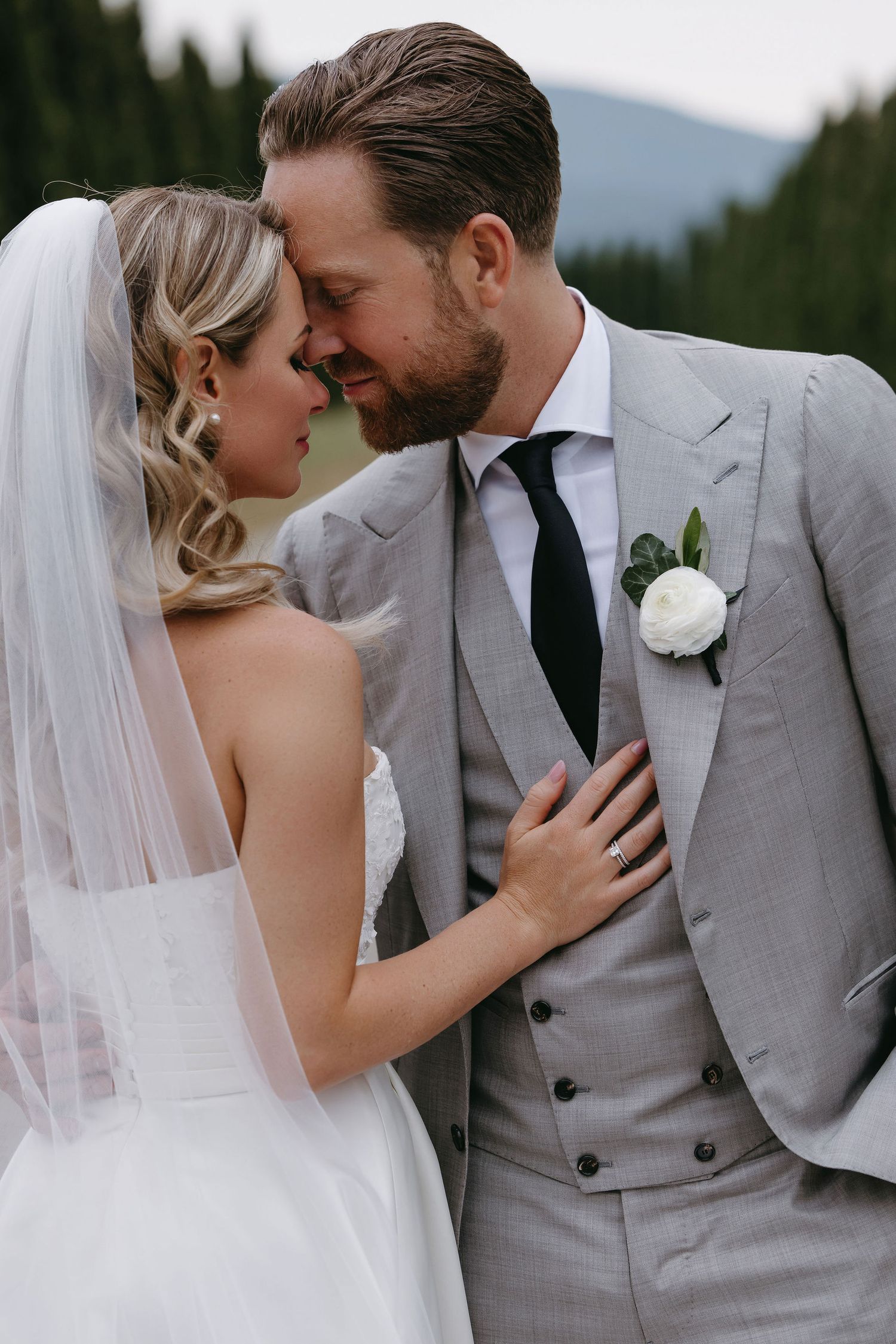 Intimate wedding portrait with couple in elegant gray suit and white dress against mountain backdrop.