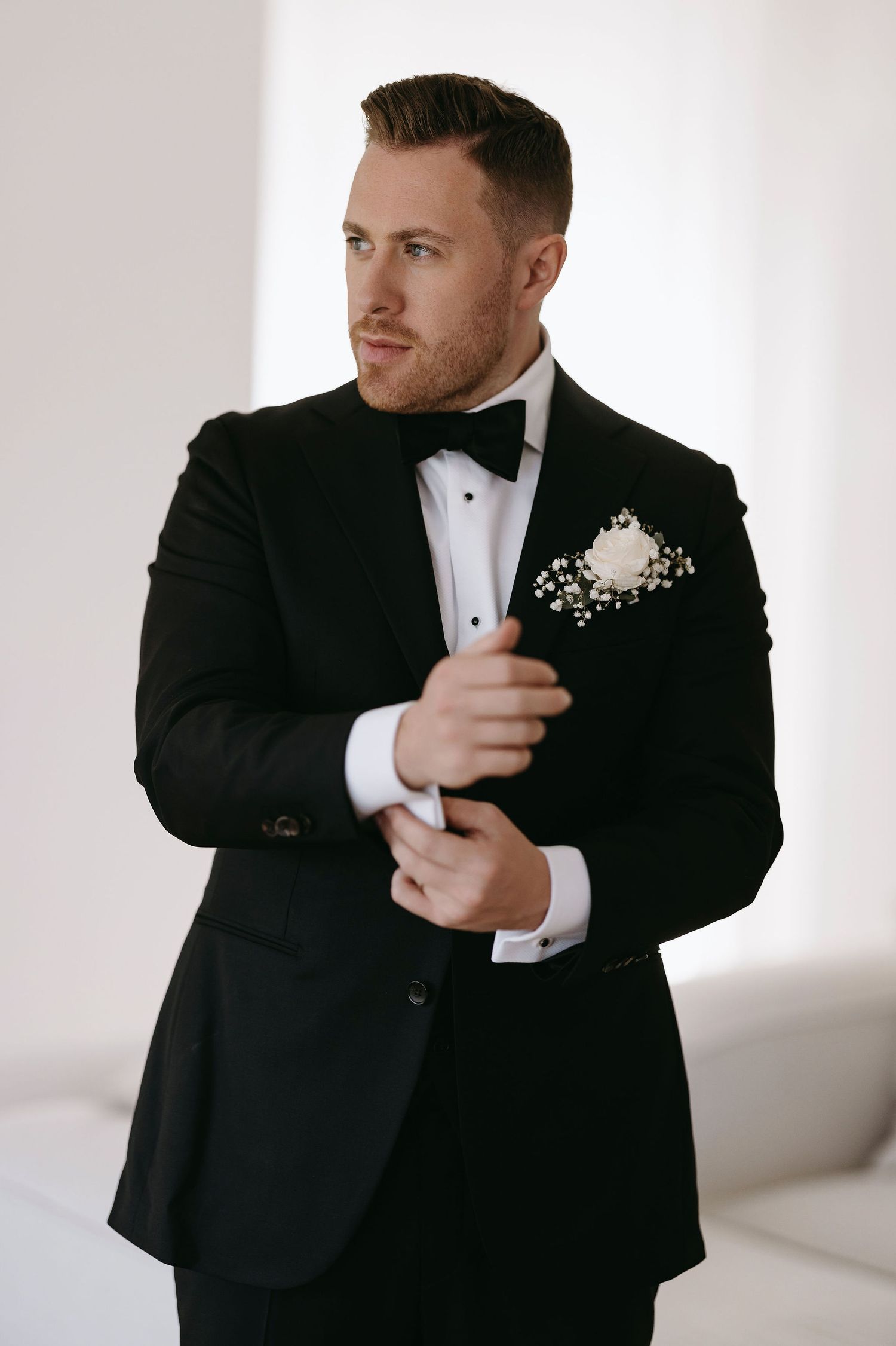 A groom in a classic black tuxedo adjusts his cuff while getting ready.