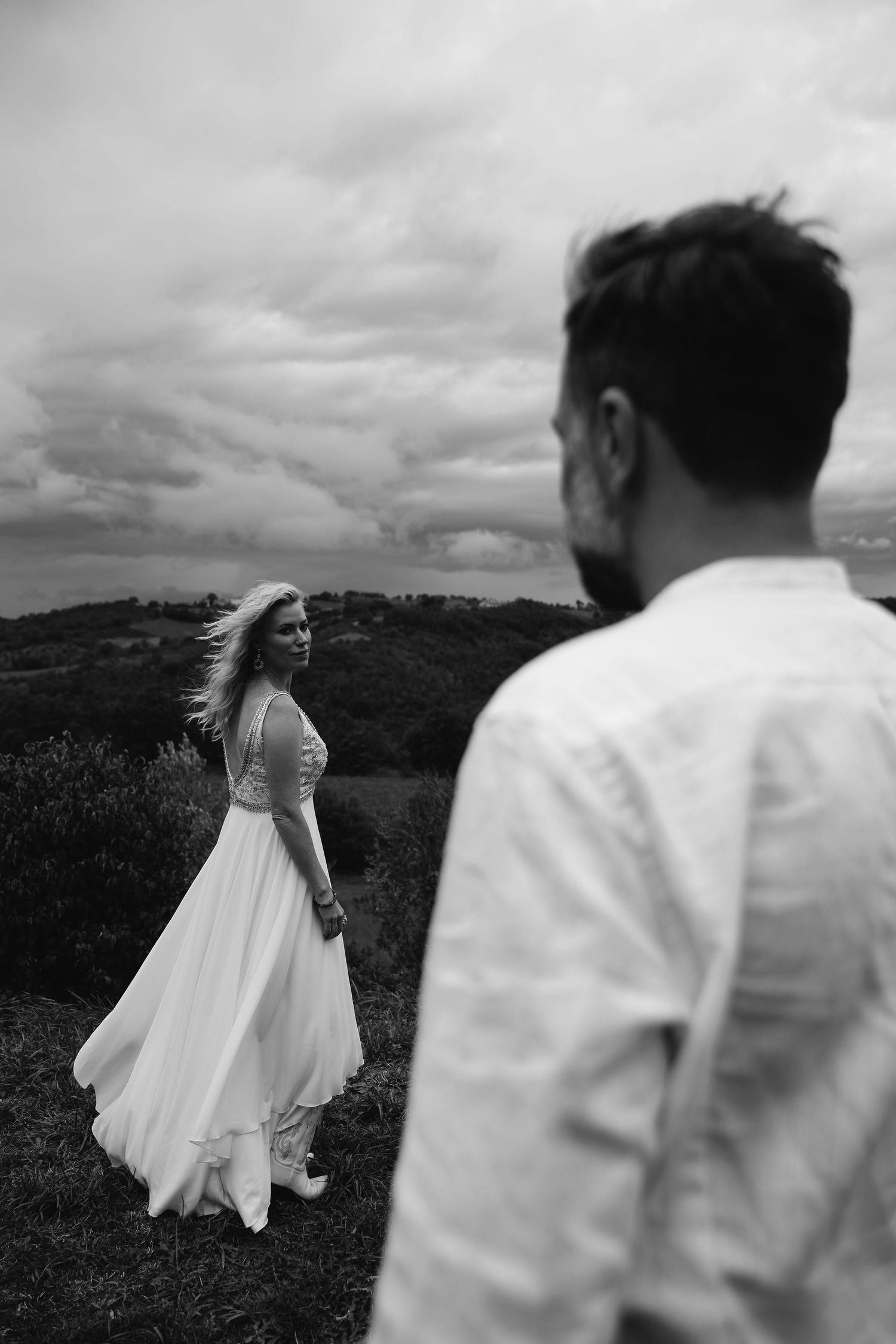 Black and white wedding photo of a couple on a hilltop, with the bride's white dress flowing in the wind.