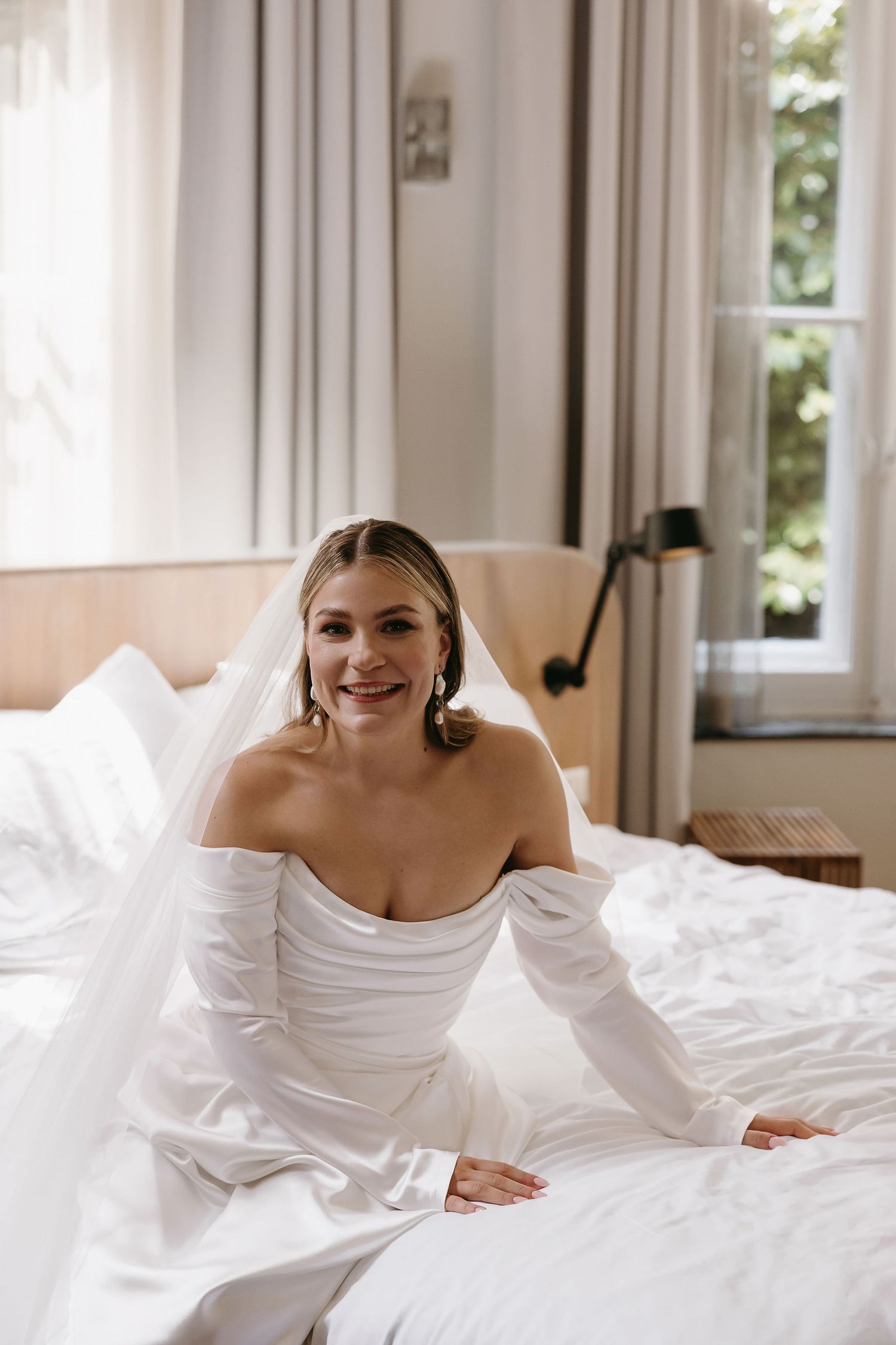 A bride wearing an elegant off-shoulder white wedding gown sits on a bed in a bright, sunny room.