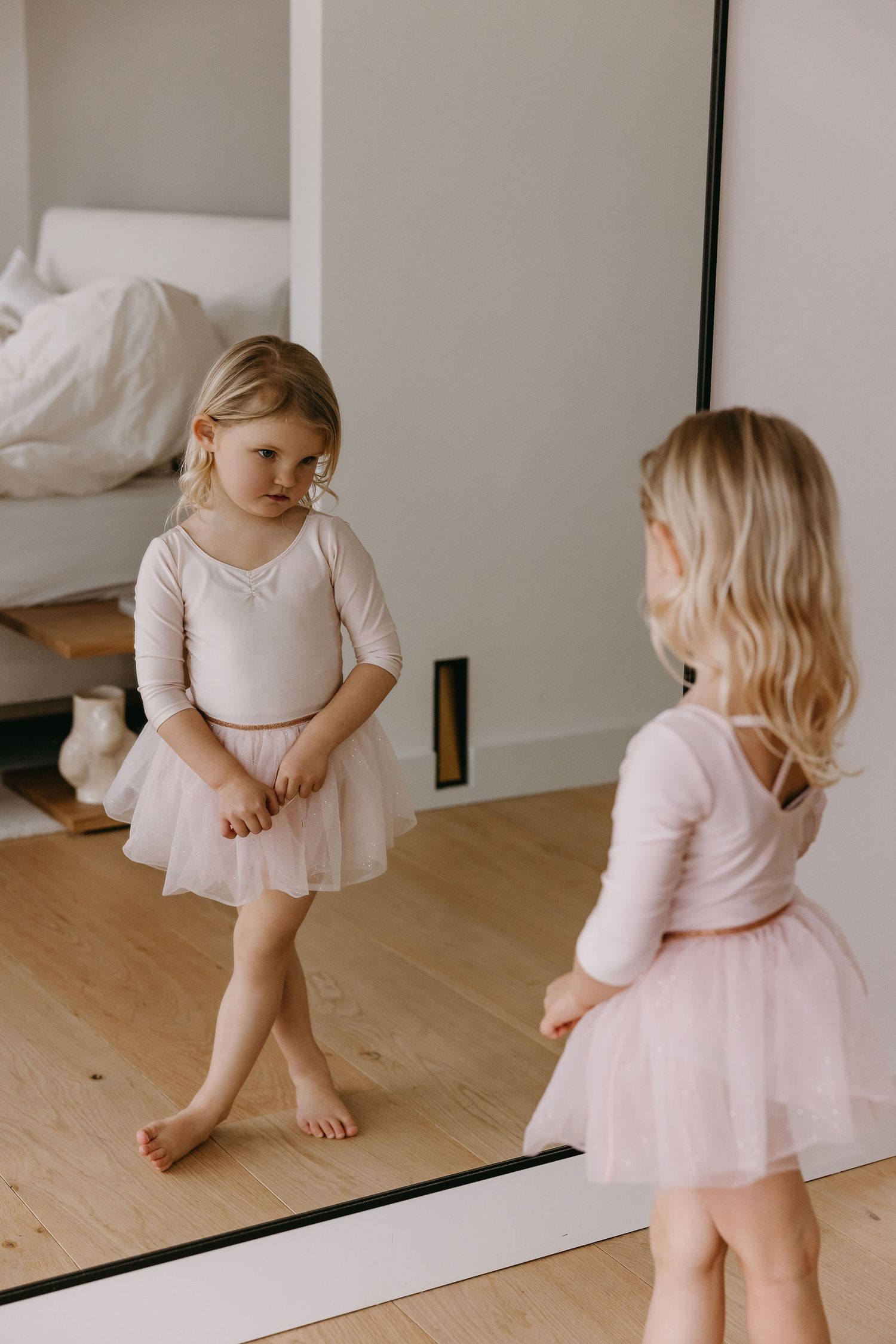 A young dancer in pink tutu looks at their reflection in a mirror.