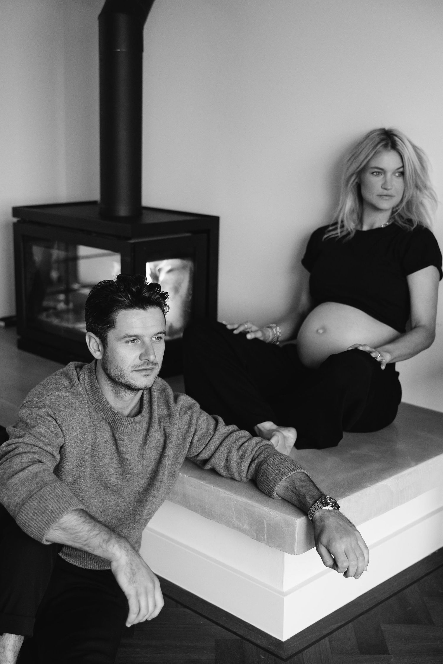 Intimate black and white maternity photo of couple relaxing by modern fireplace.