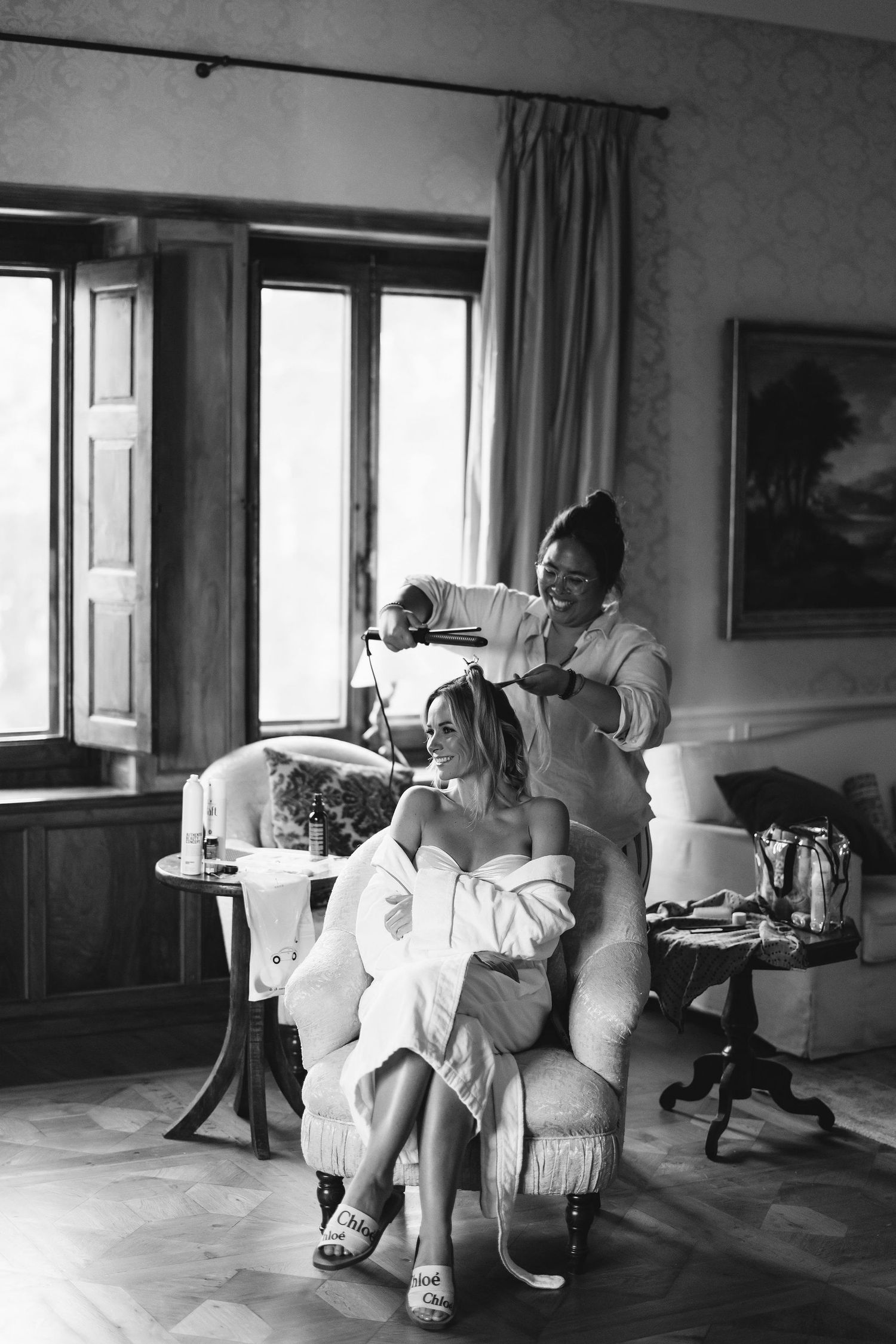 Black and white image of a hair styling session in an elegant room with large windows.