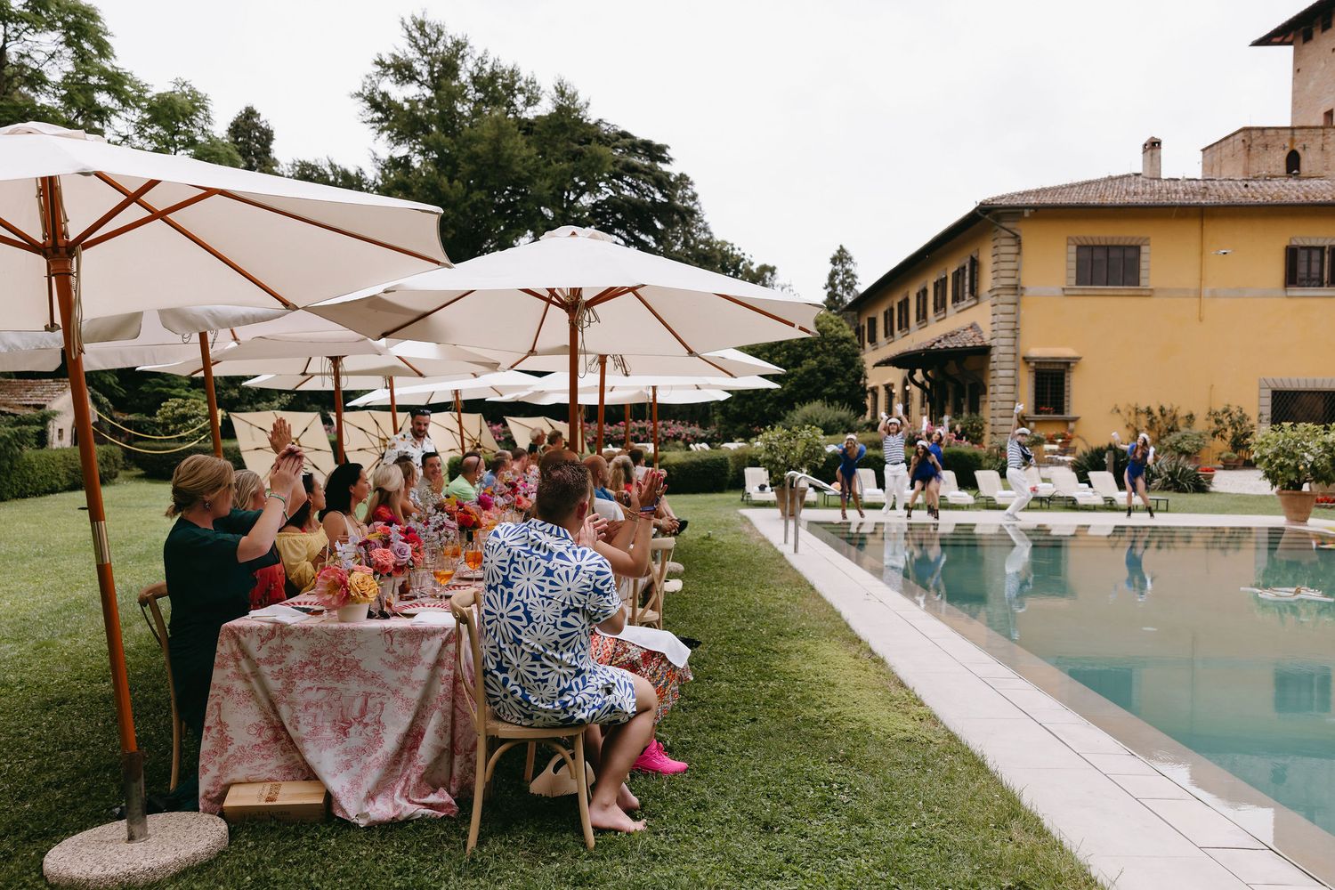 Wedding reception by a luxurious pool with guests dining under white umbrellas at a Tuscan villa.