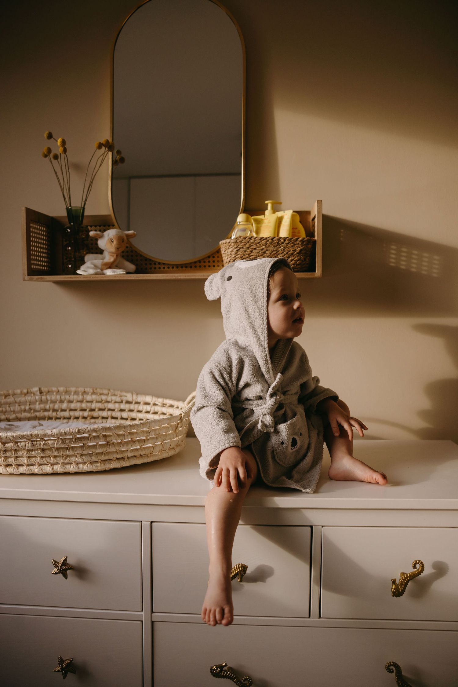 A small child in a hooded outfit sits on a white dresser near an arched mirror.