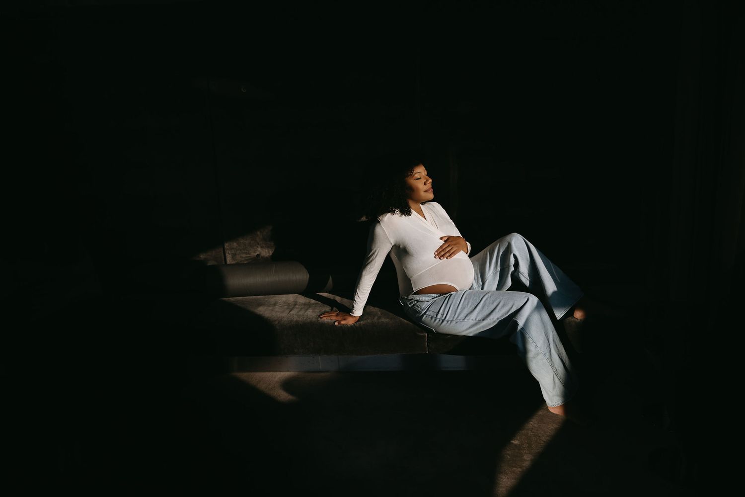 Person in white clothing reclines on dark sofa in moody lighting.