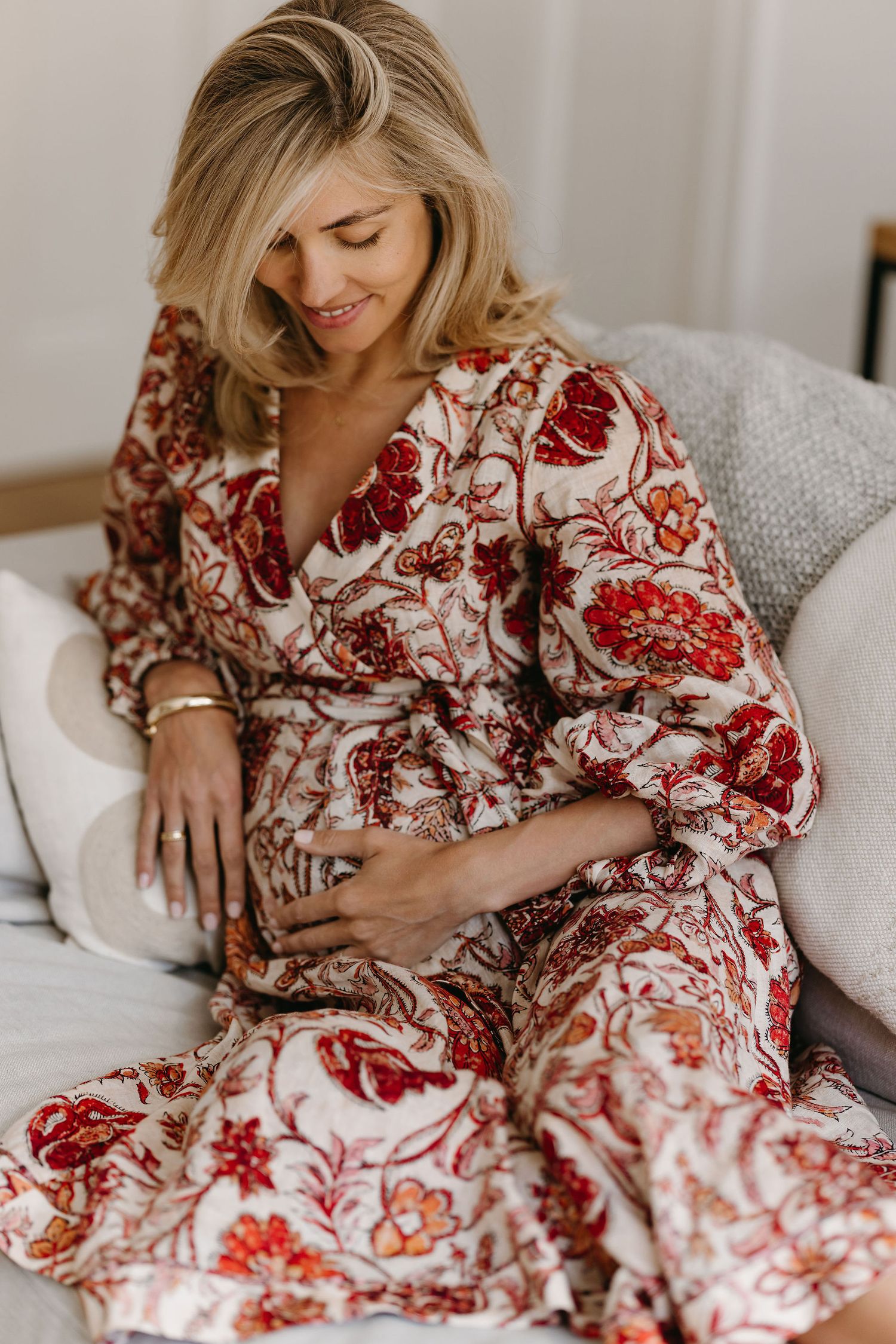 Expectant mother in red and white floral dress relaxes on couch during maternity photoshoot.