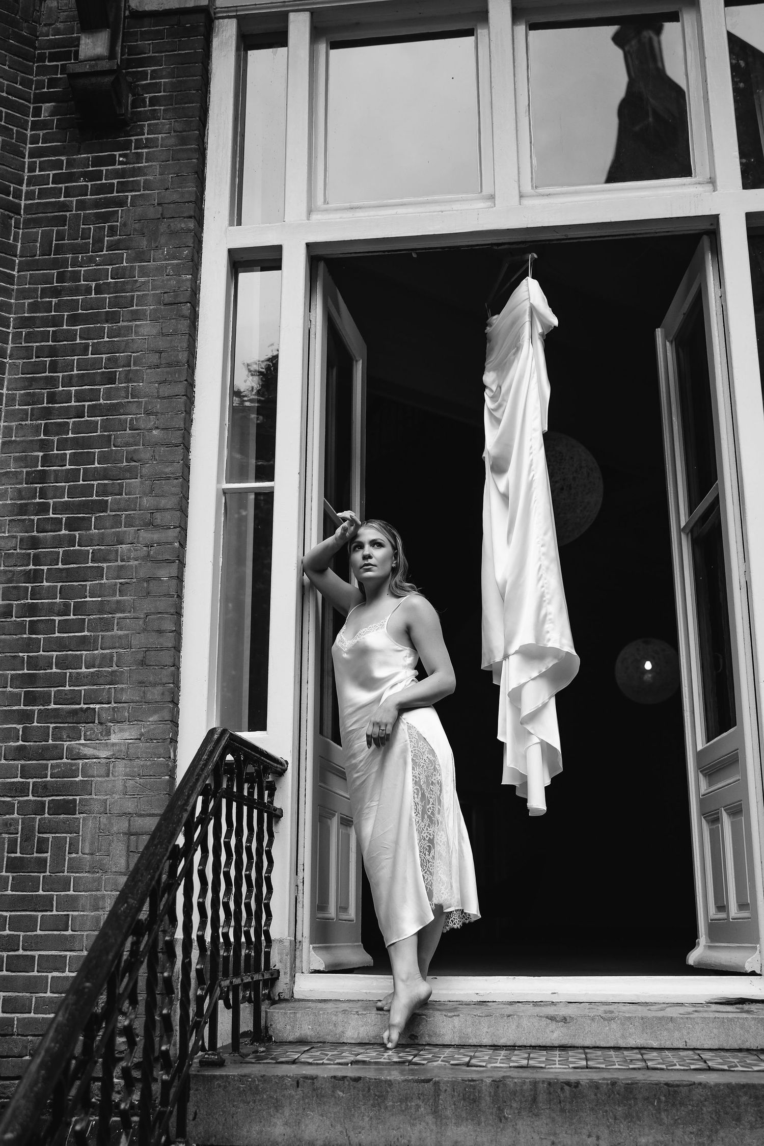 Black and white portrait of bride standing in doorway with dress hanging above.