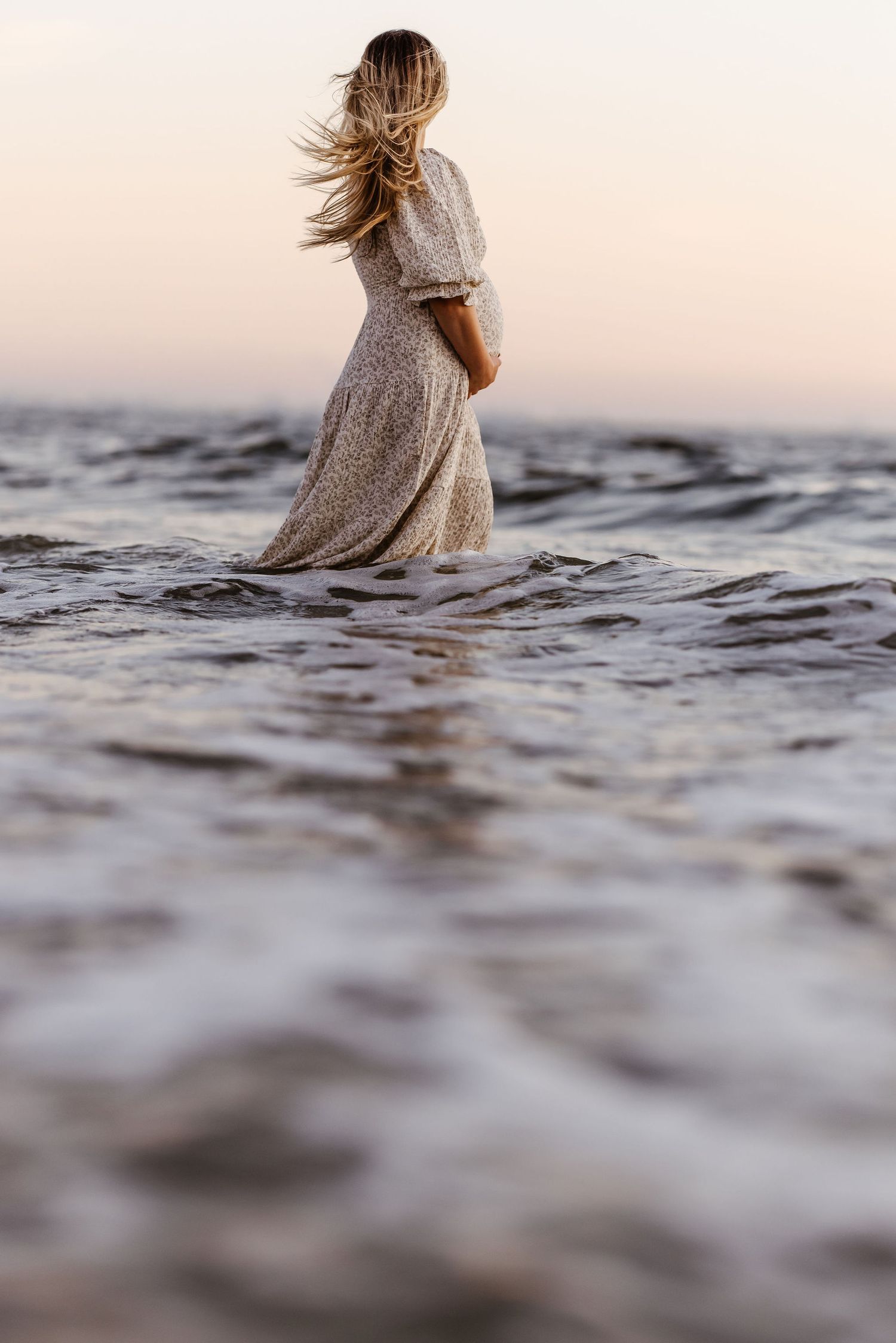Woman in a flowy dress stands in shallow ocean water at sunset with hair blowing in the wind.