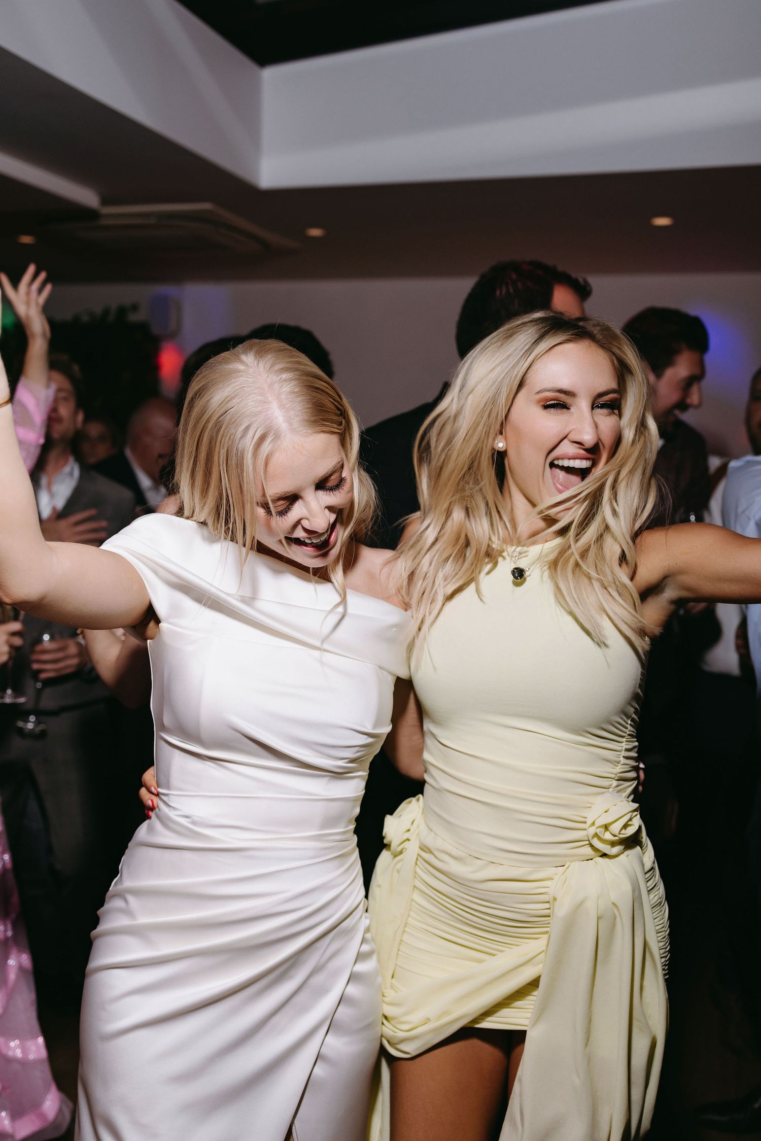 Two women in light-colored dresses dancing and celebrating at a wedding reception.