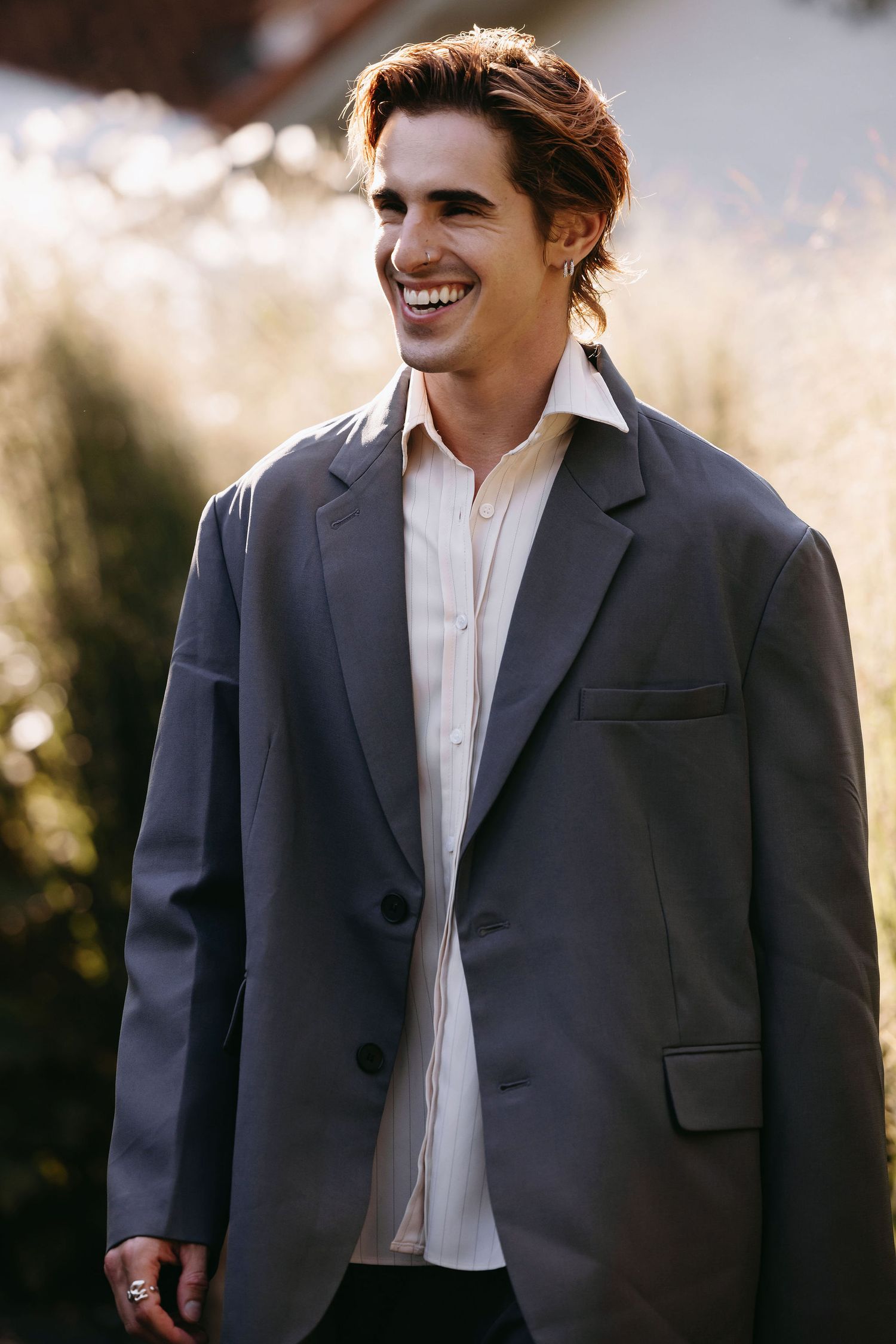 Young man in dark blazer and white shirt smiles outdoors with warm sunlight.