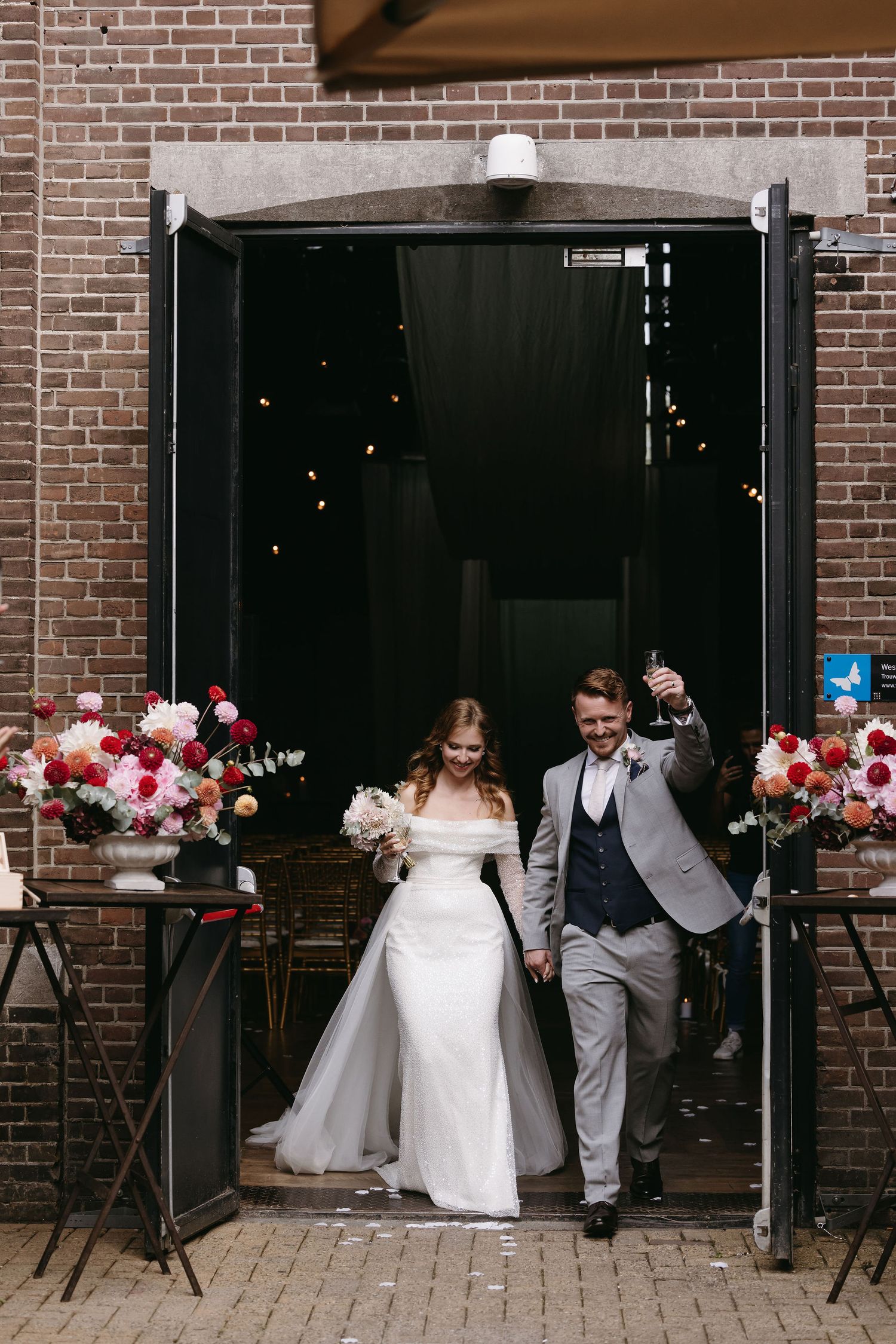 Couple exits through brick doorway decorated with pink and red floral arrangements at wedding venue.