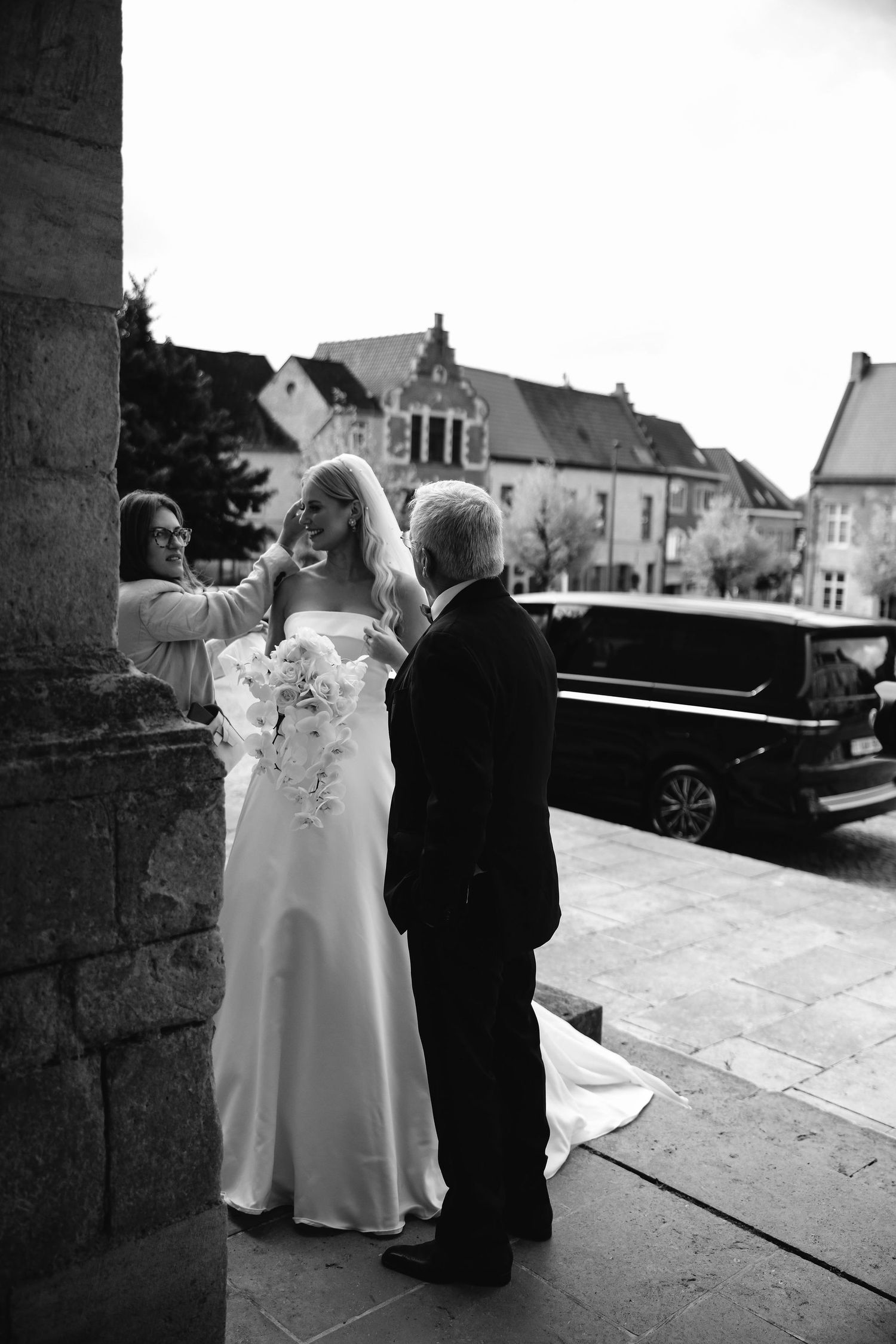 Wedding party preparing outside historic stone church with vintage car in background.