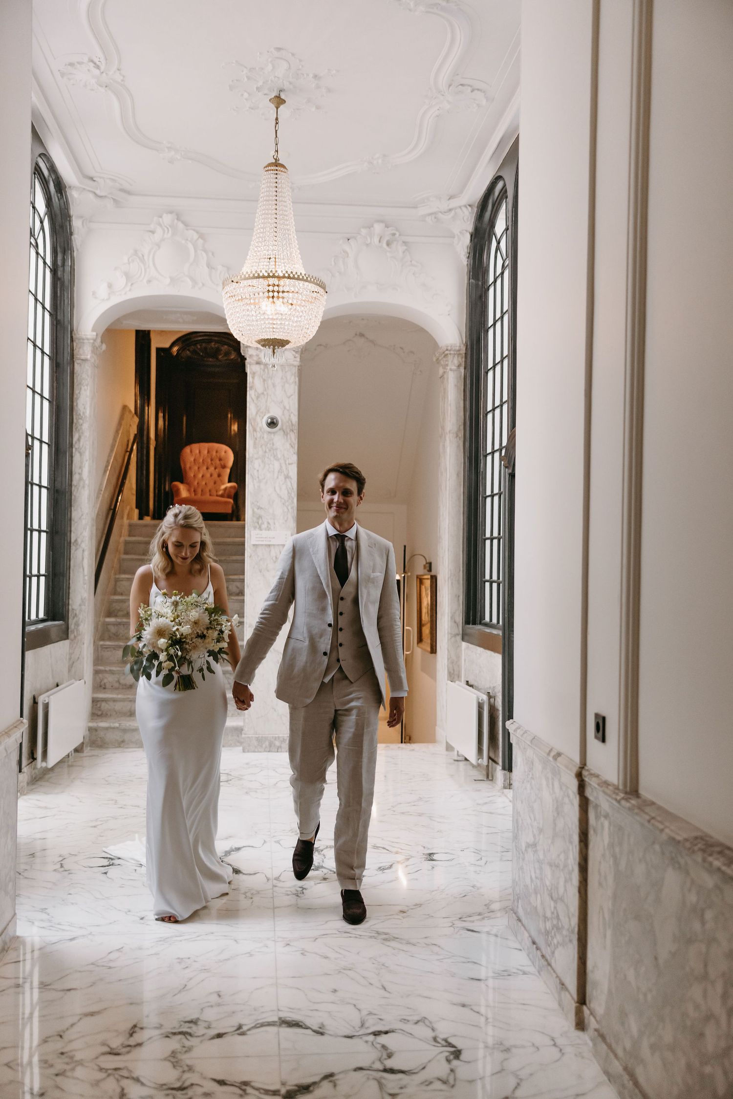 Couple running playfully through an elegant marble hallway with crystal chandeliers.