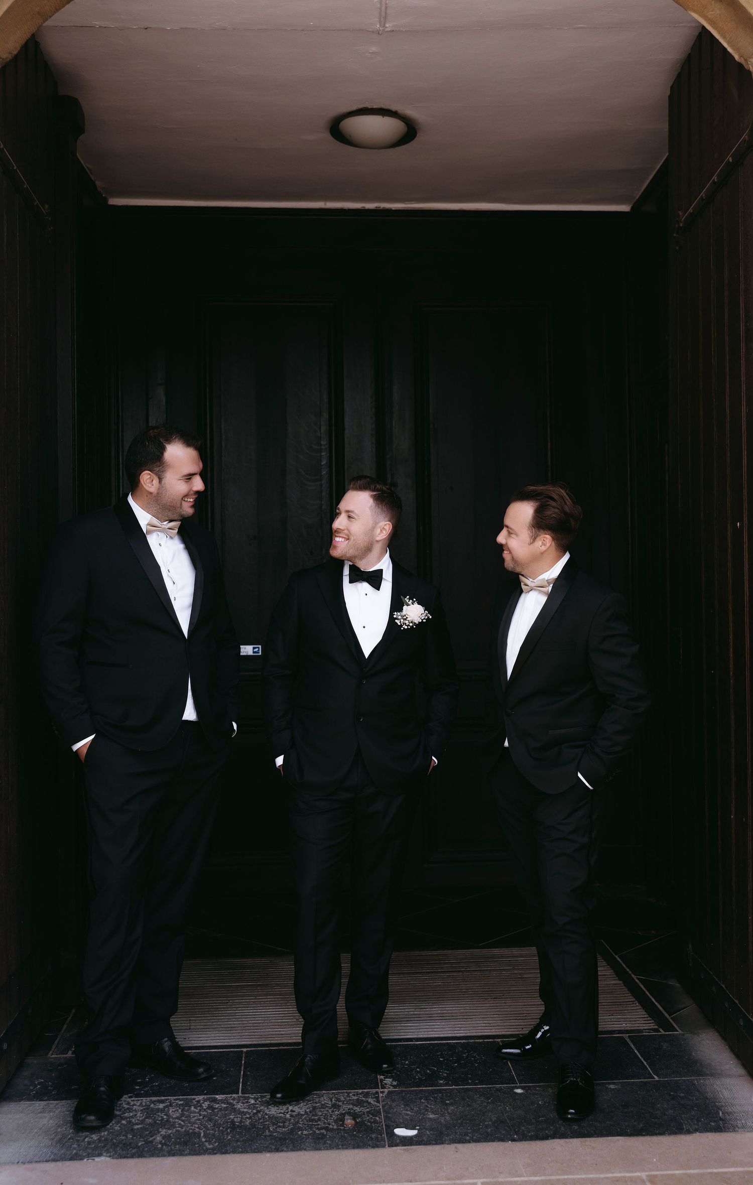 Three groomsmen in formal tuxedos share a candid moment in dark doorway.