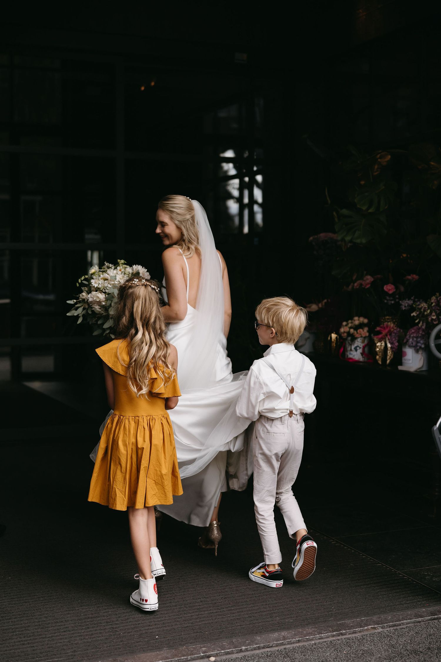 Two children in white and yellow outfits follow a person in a white dress.