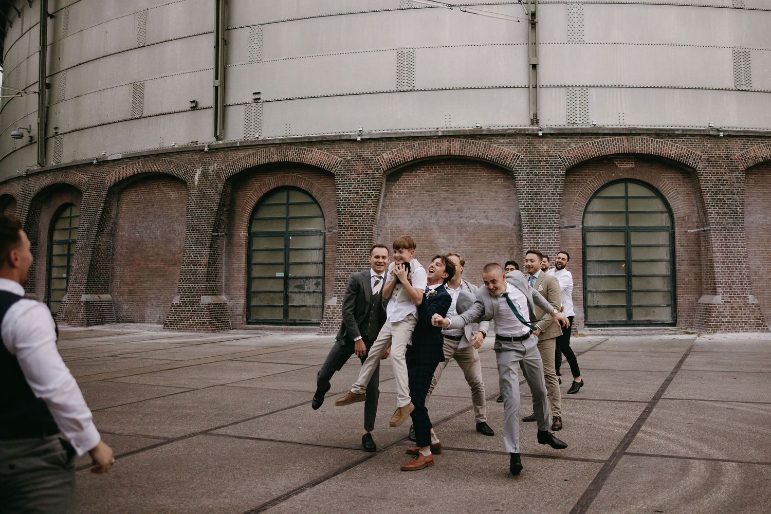 Wedding party members playfully dance and pose outside historic brick building.