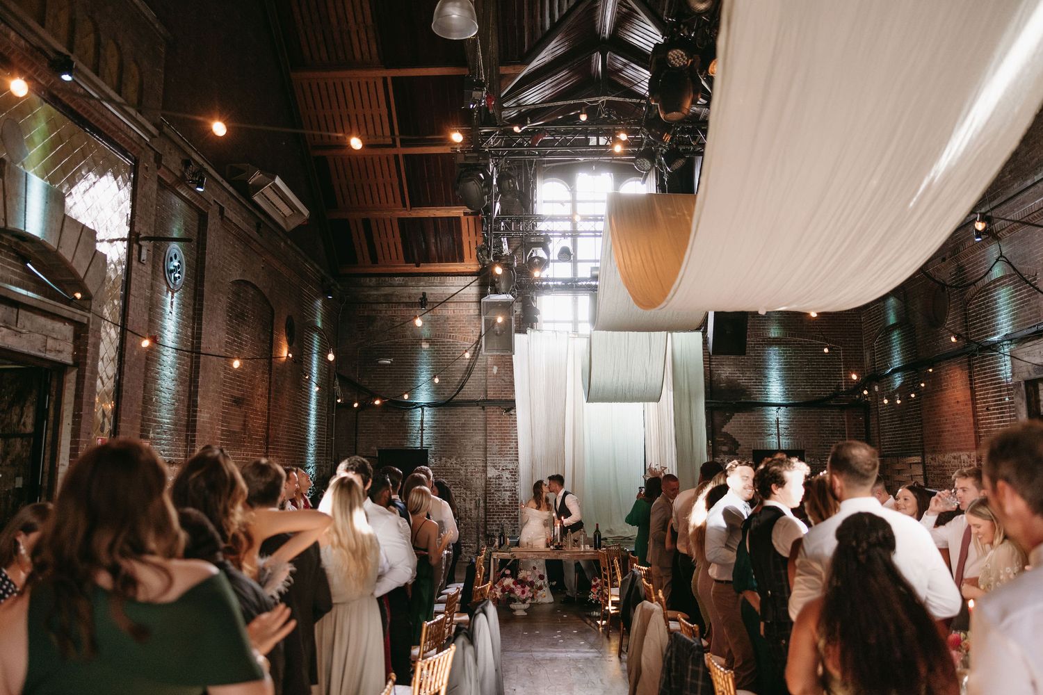 Wedding reception in a dimly lit industrial venue with exposed brick walls and hanging lights.
