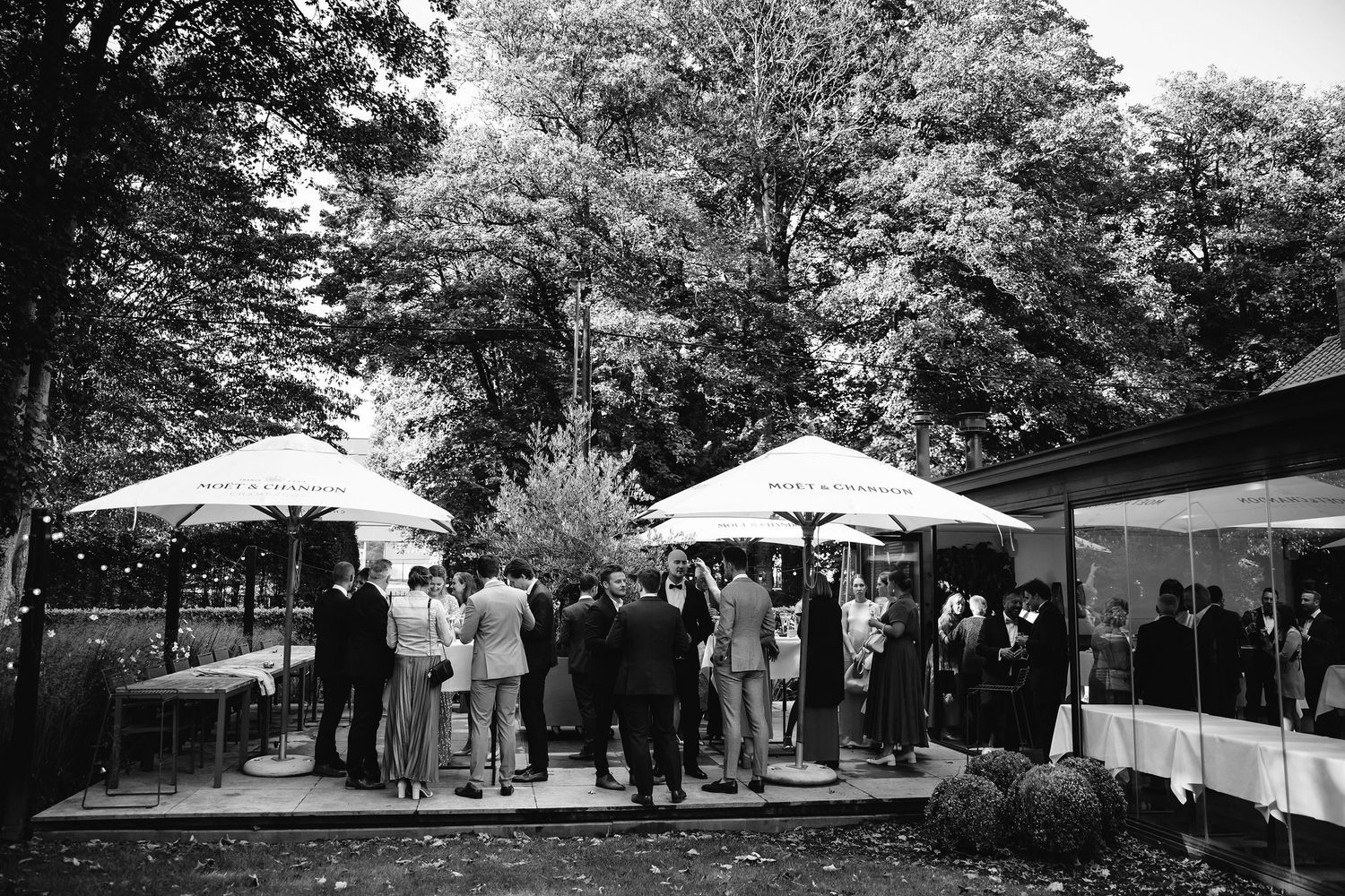 Wedding guests gather under white umbrellas at an outdoor reception venue surrounded by trees.
