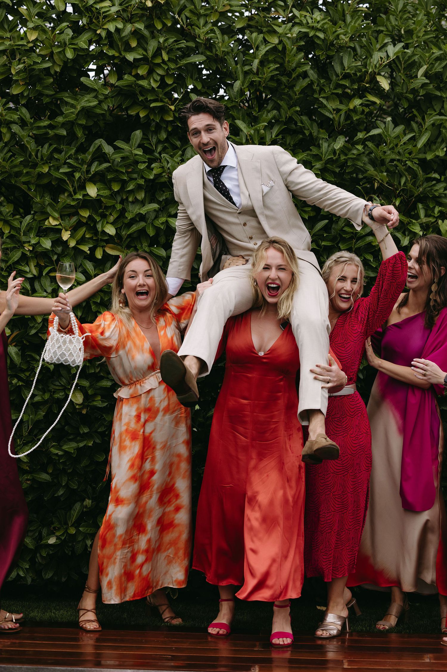 Celebratory wedding party photo with people in vibrant dresses jumping and laughing.