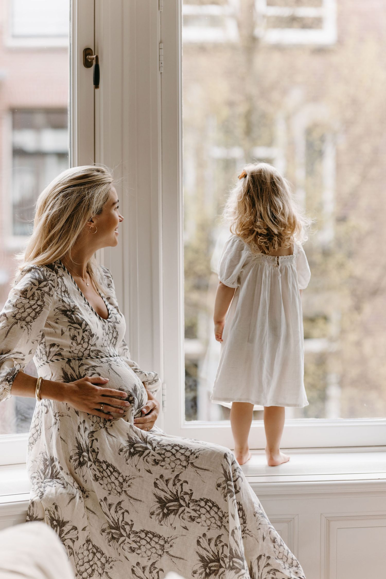Maternity photo with floral dress and white dress by sunlit window.
