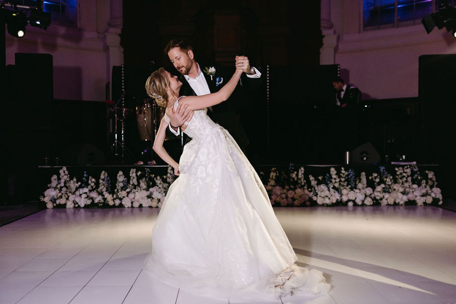 A couple performs their first dance in an elegant ballroom with a white dance floor and ambient lighting.