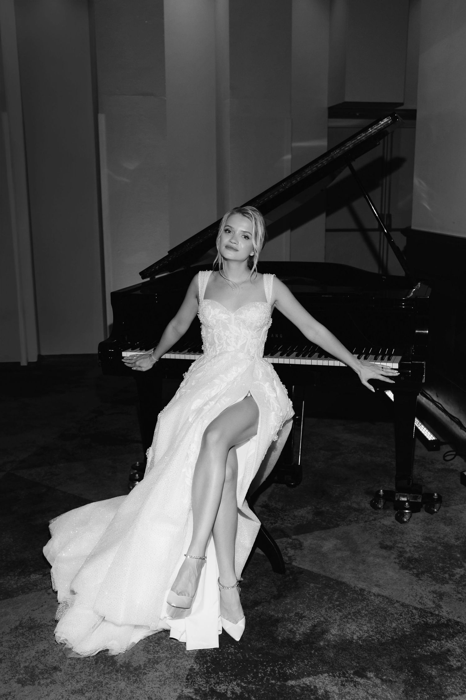 Black and white portrait of a bride in a flowing dress seated at a grand piano in an elegant performance space.