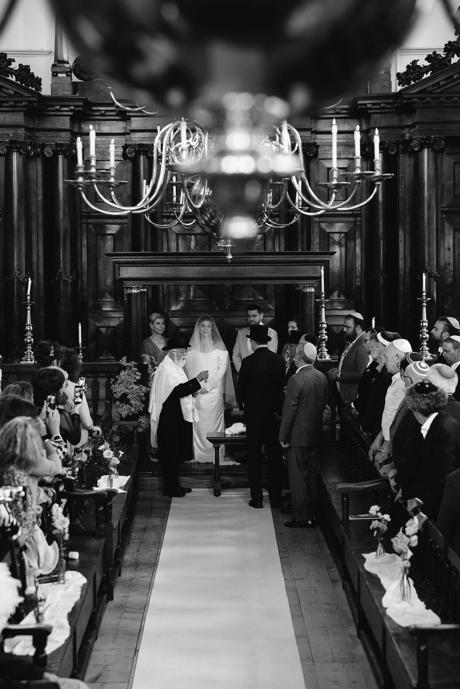 Black and white photograph of a traditional church wedding ceremony with guests seated in pews under an elegant chandelier.