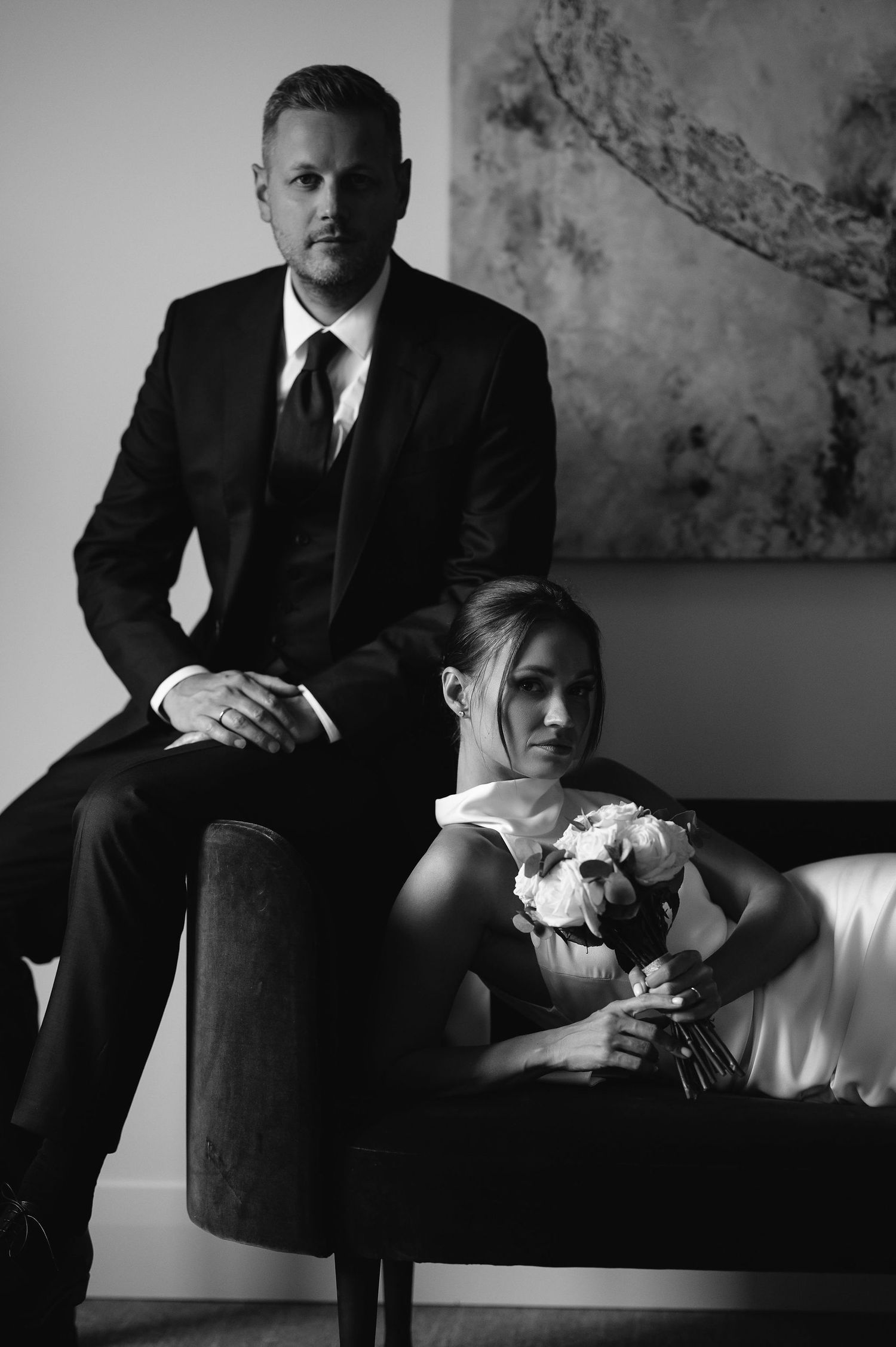 A black and white wedding portrait shows a couple posing with a bouquet on an elegant couch.