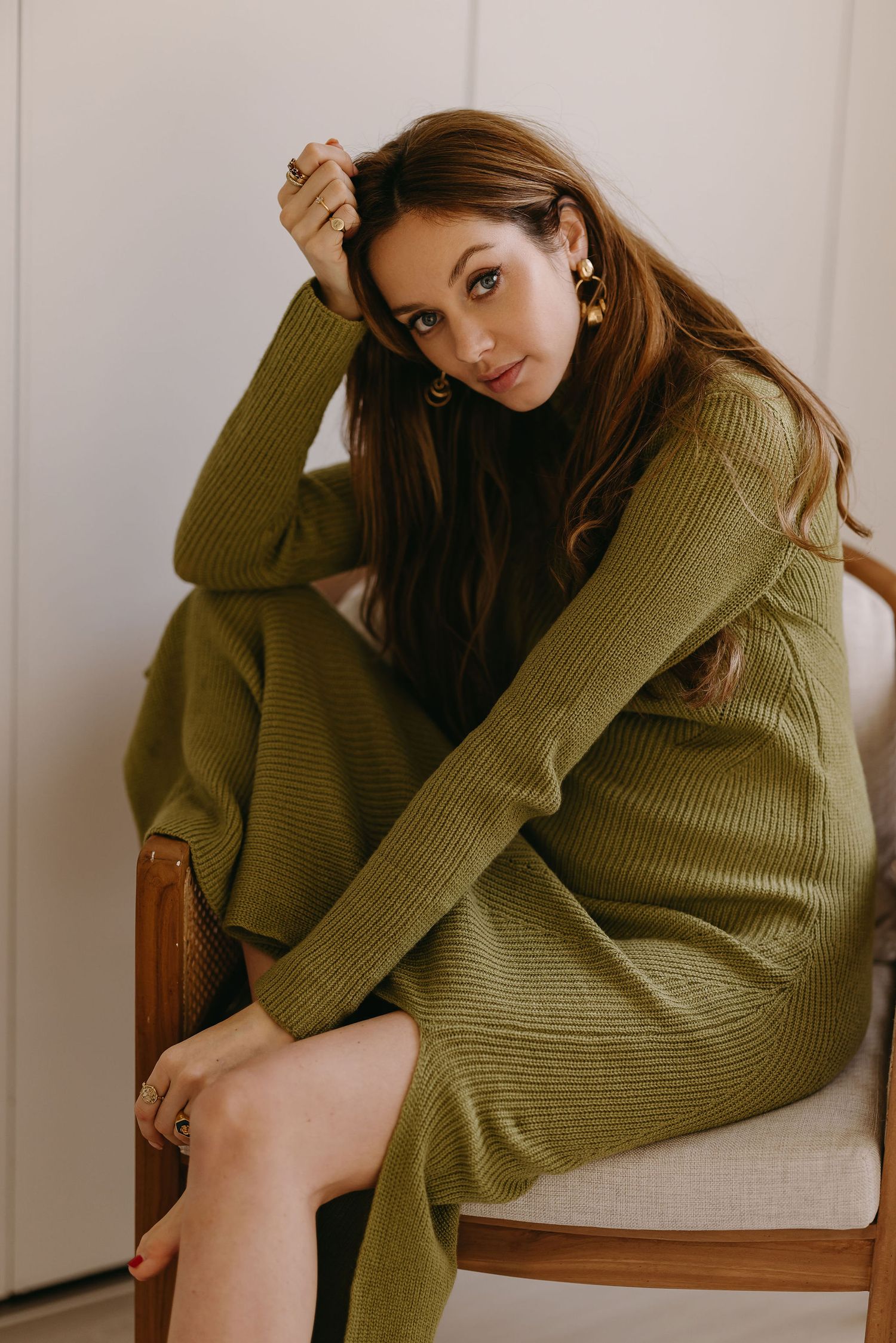 Person in olive green sweater lounging pensively in wooden chair by window.