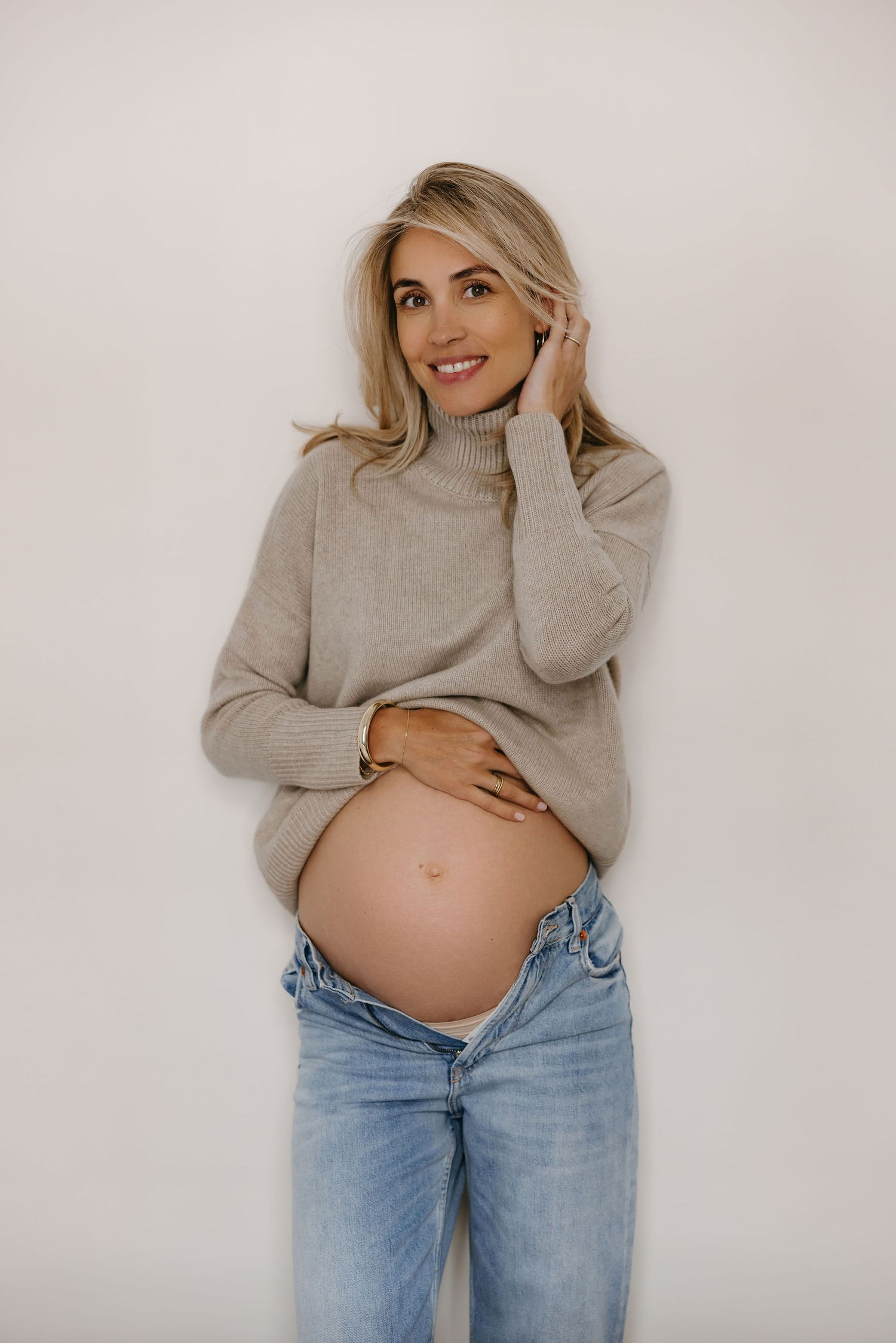 Expecting mother in grey sweater and jeans poses against white backdrop showing pregnancy journey.