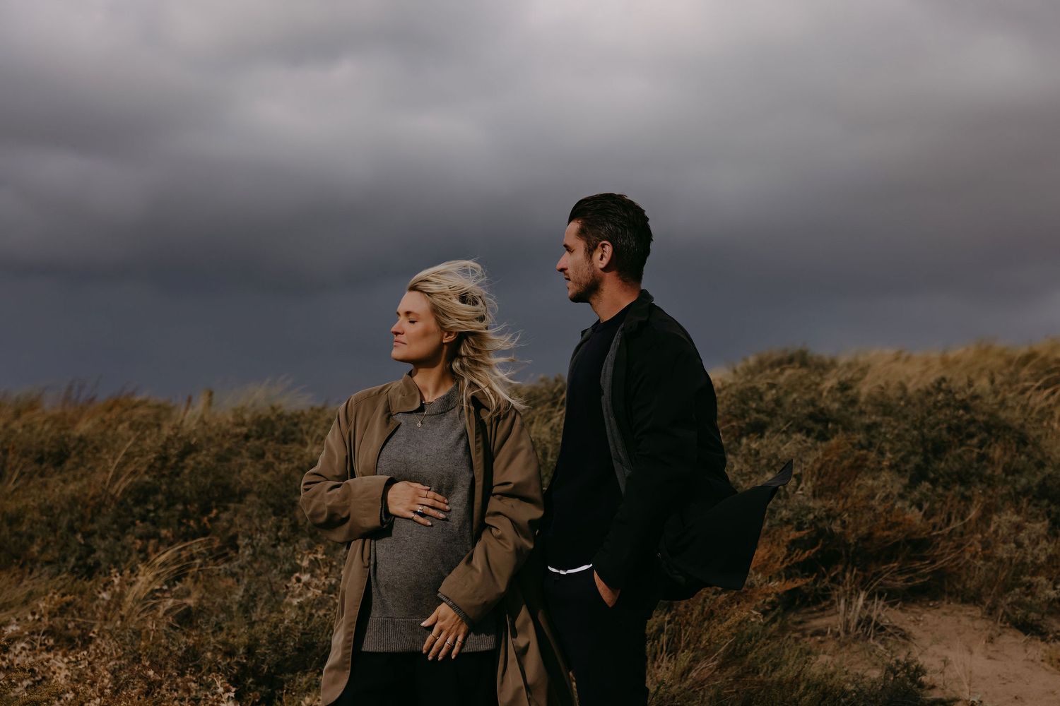 Intimate outdoor maternity photo showing couple embracing against moody sky and coastal landscape.