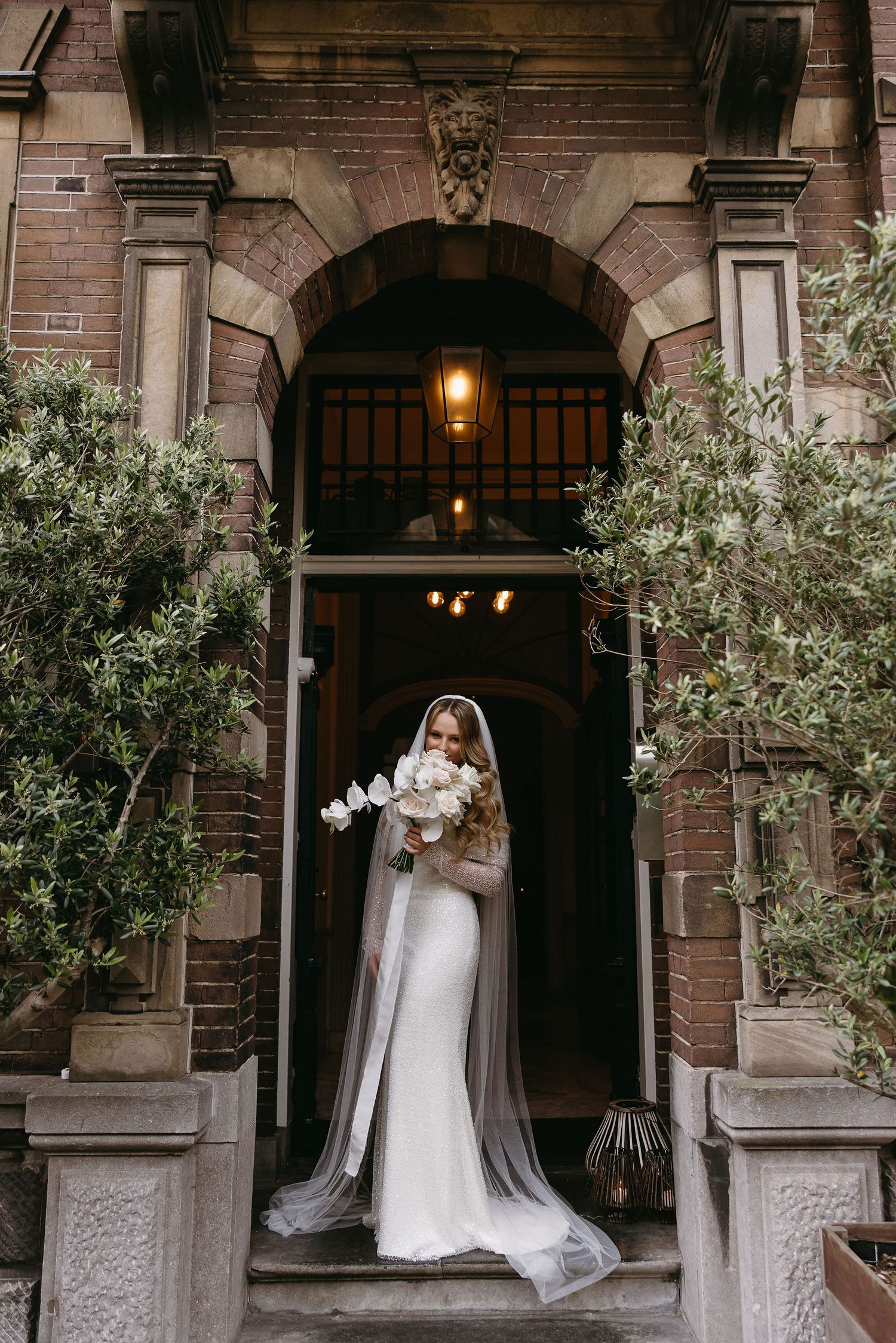 Bride in fitted white gown with cascading bouquet poses in ornate stone doorway framed by olive trees.