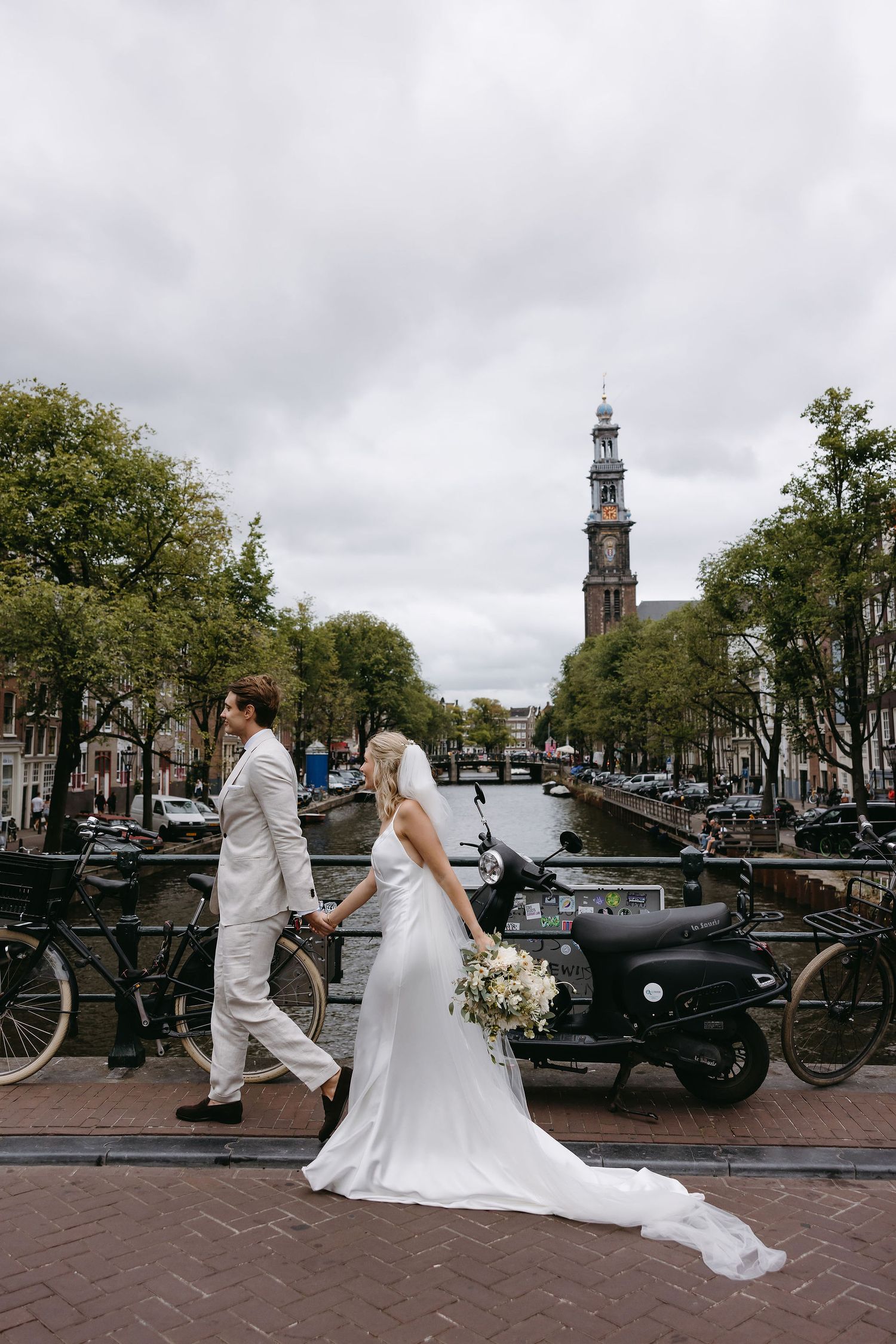 Wedding couple walking along Amsterdam canal with church spire and bikes in background.