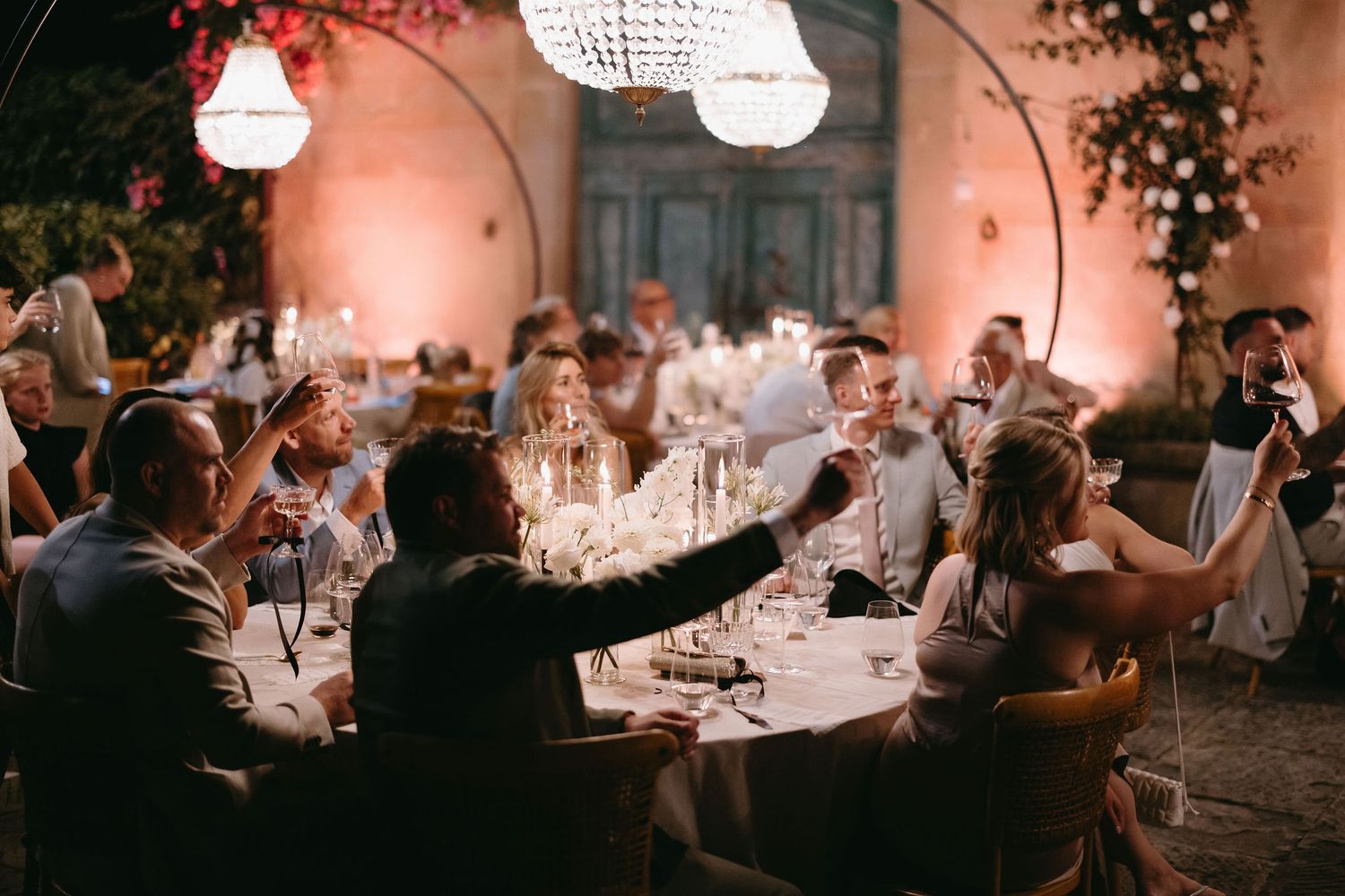 Evening wedding dinner party with guests toasting under string lights and chandeliers.