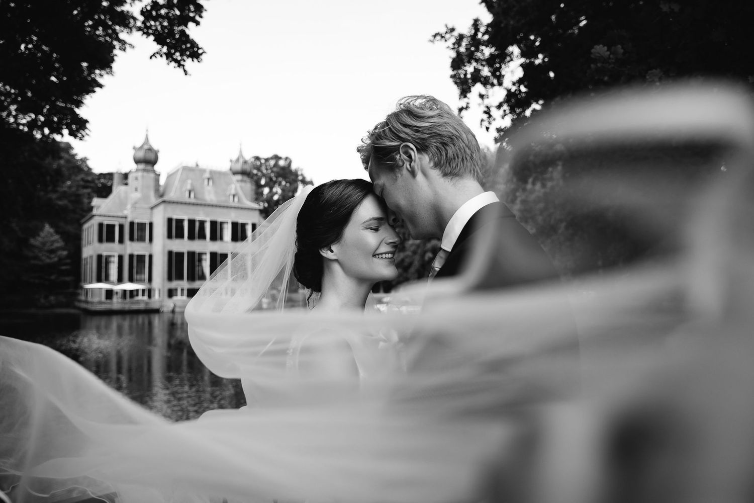A romantic black and white portrait of newlyweds with a grand estate and lake in the background.