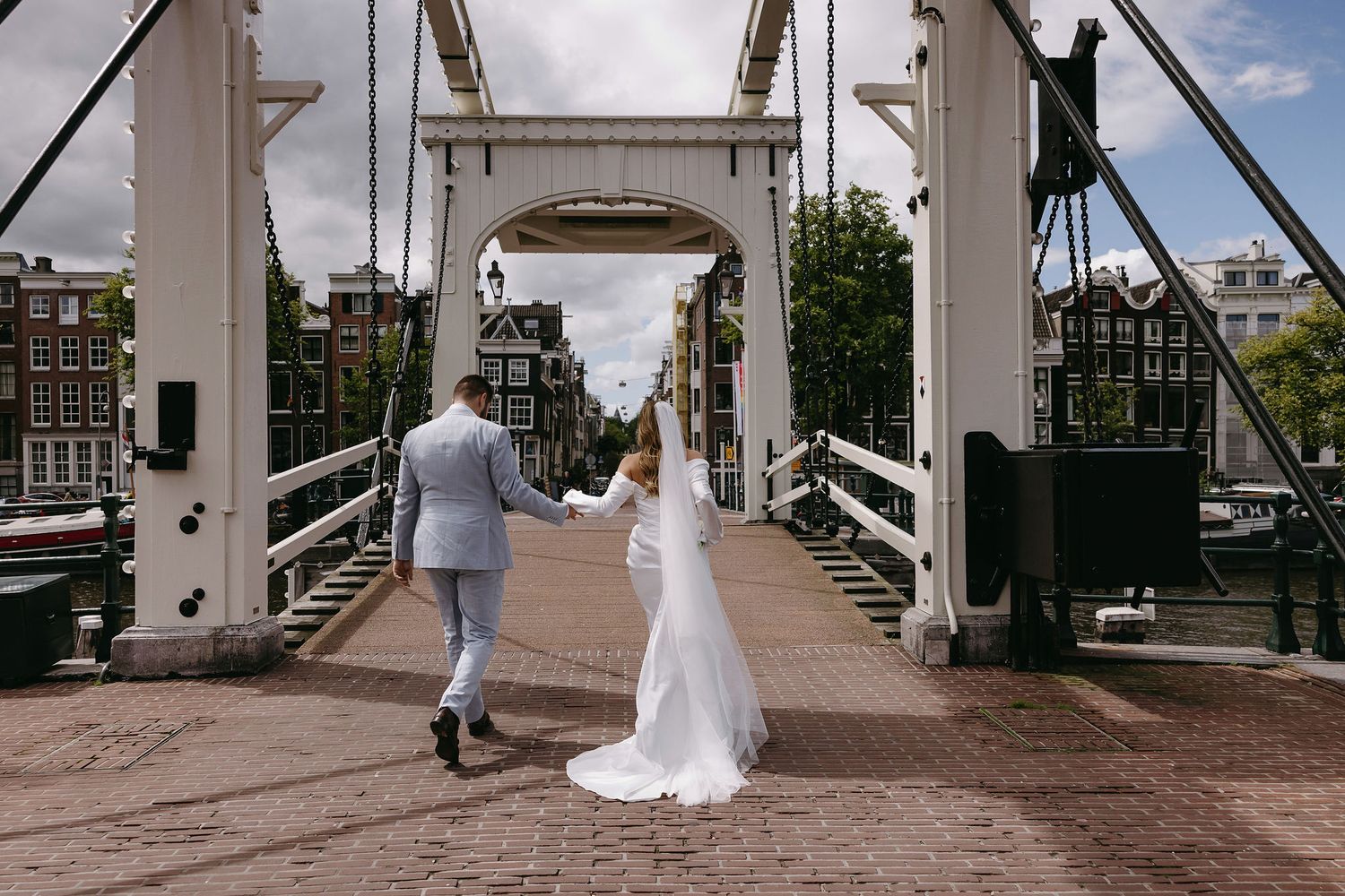 A couple walks across a historic white bridge in wedding attire.