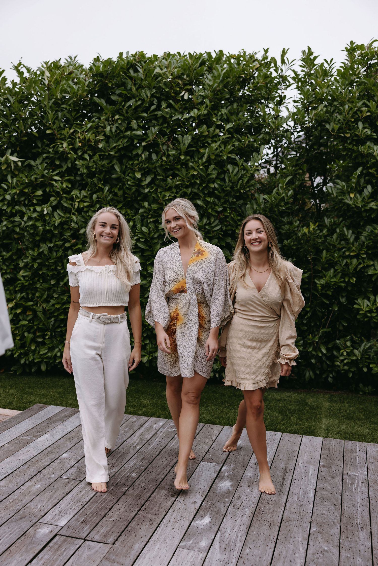 Three women in neutral toned dresses and outfits walk together on a wooden deck.