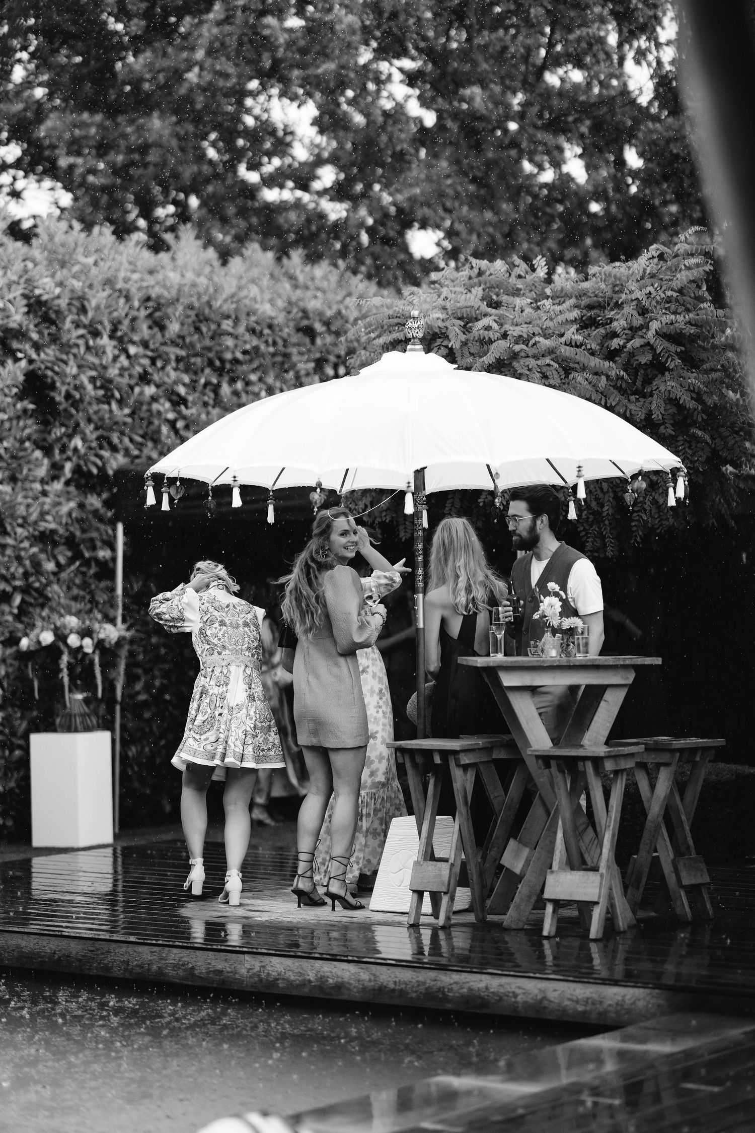 Guests gather around tables under a white umbrella at an outdoor garden party in black and white vintage style.